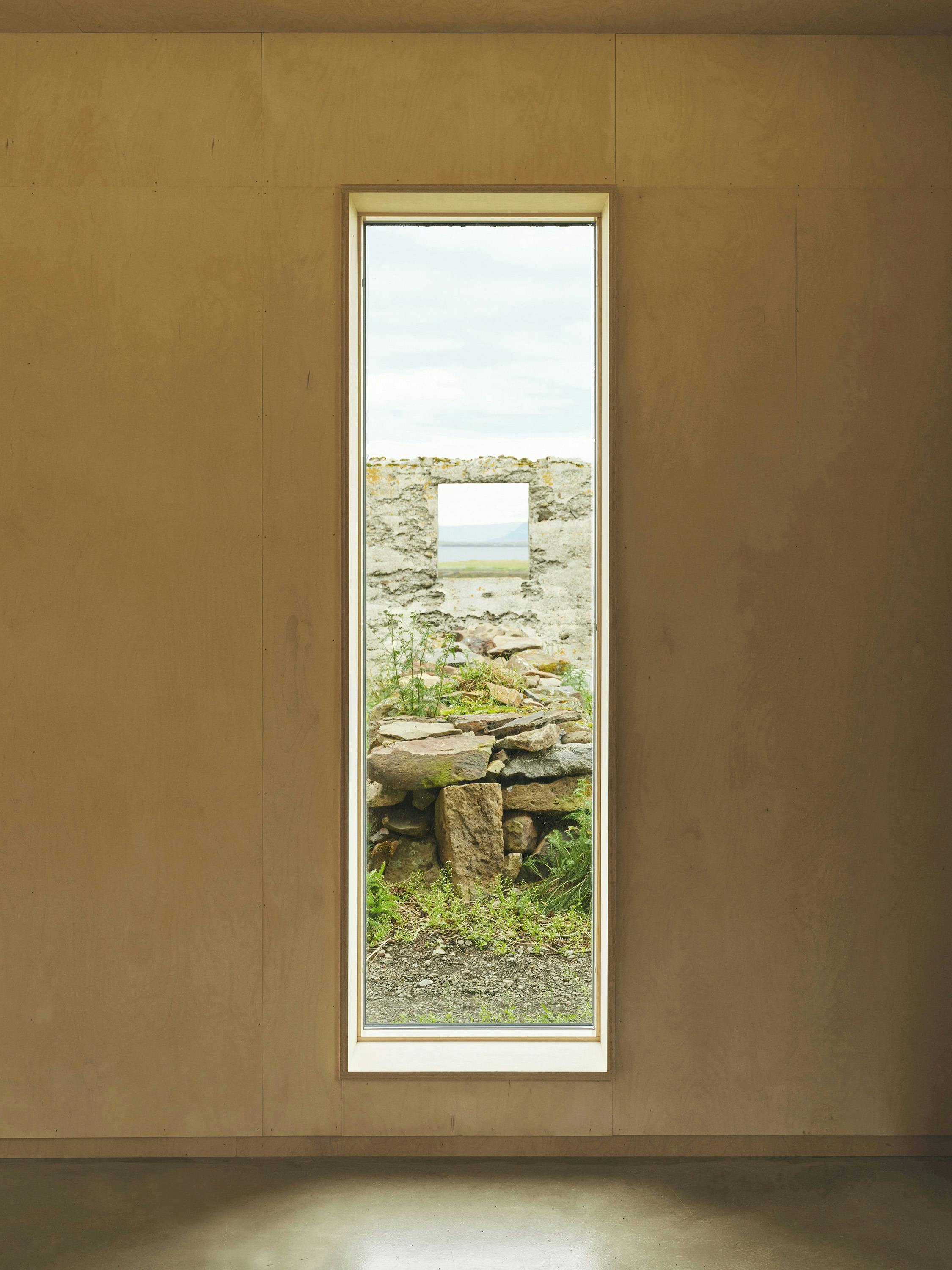A straight-on shot of a rectangular window framed by a light-colored wooden wall. The window reveals an outdoor scene of a dilapidated stone structure with a rectangular opening that mirrors the shape of the window itself. The stone structure appears to be a ruin, with weathered stones and patches of green grass and moss growing within and around it. Through the opening in the stone structure, a glimpse of a body of water and a cloudy sky is visible. The wooden wall surrounding the window is smooth and light-colored, with a subtle texture. The floor beneath the window is a darker shade, with a slight shadow cast across it. The lighting is soft and diffused, creating a serene and contemplative atmosphere. The image conveys a sense of architectural framing and the juxtaposition of interior and exterior spaces.