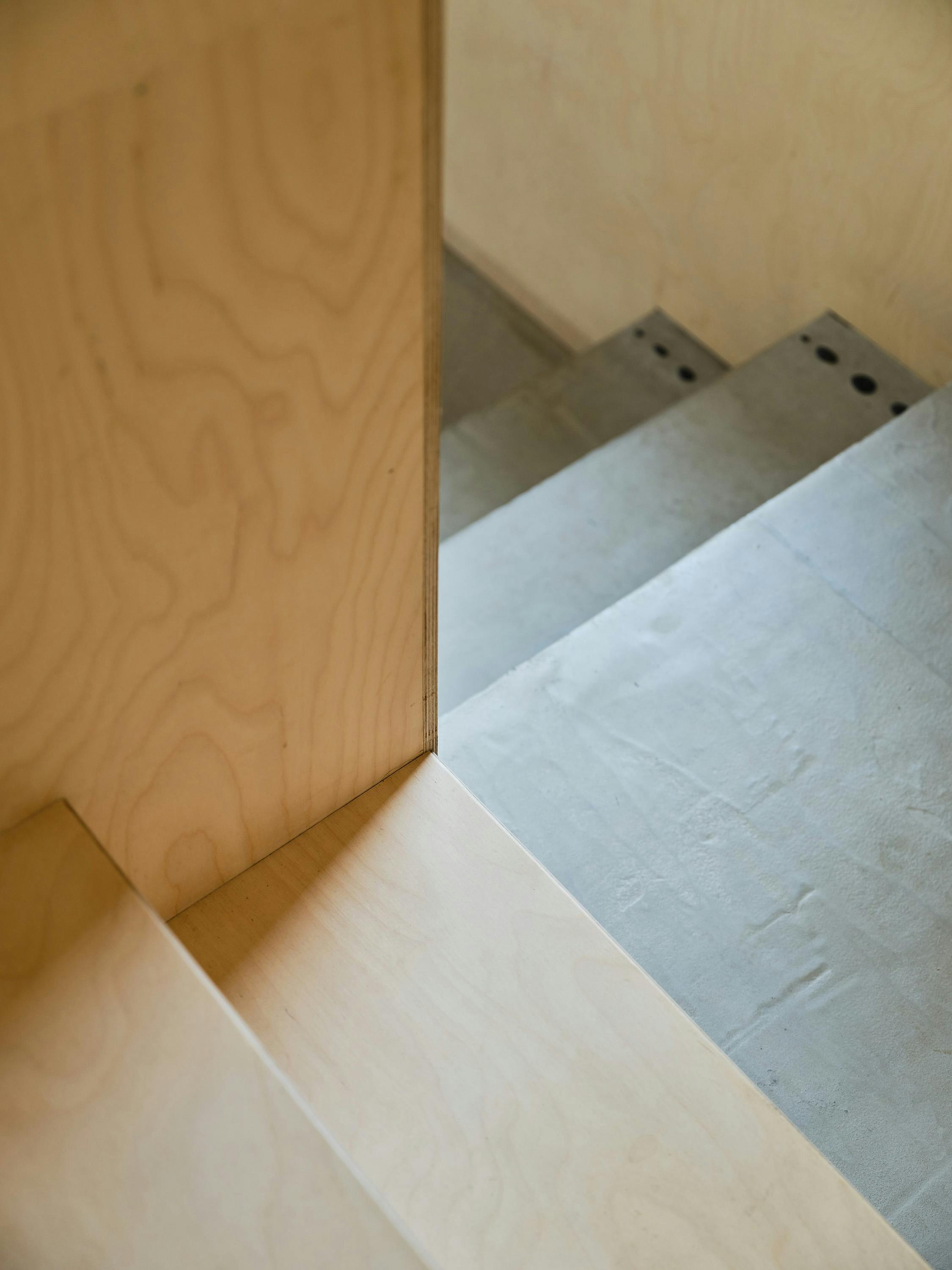 A close-up, slightly angled photograph of a staircase detail. The image focuses on the junction between a light-coloured wood panel and concrete steps. The wood panel has a visible grain pattern and a clean, straight edge. The concrete steps are smooth and light grey, with small, dark circular indentations visible on the risers. The perspective is from above, looking down the staircase. The lighting is soft and natural, highlighting the textures and materials. The overall aesthetic is minimalist and modern, with a focus on simple, natural materials.