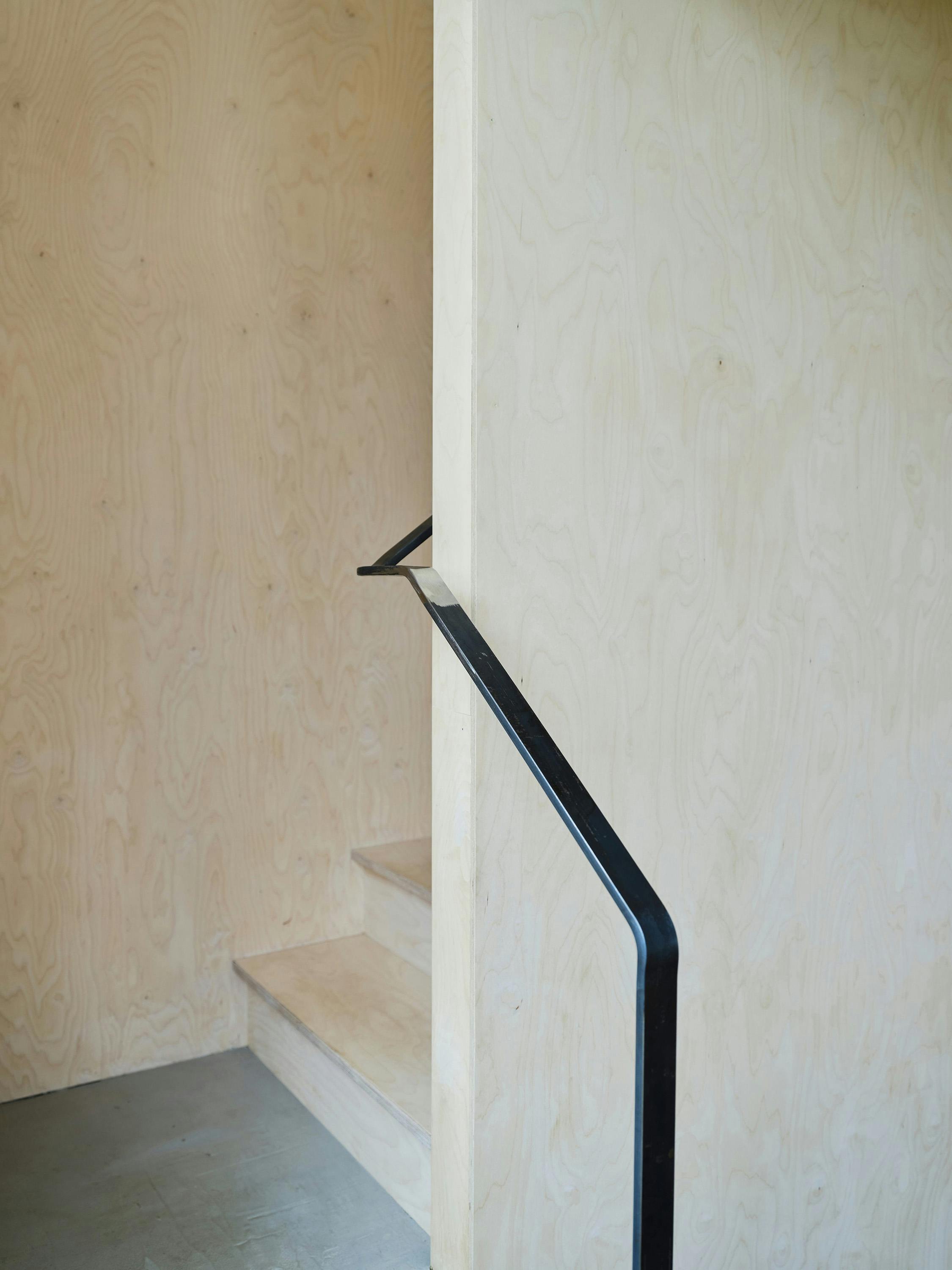 A slightly angled, eye-level photograph of a staircase detail. The image focuses on the junction between light-coloured wood panelling on the walls and the steps, and a black metal handrail. The wood panelling has a distinct grain pattern. The steps are also made of light-coloured wood. The black metal handrail is angled, running diagonally up the staircase. The floor at the bottom of the stairs is concrete. The lighting is soft and natural, highlighting the textures and materials. The overall aesthetic is minimalist and modern, with a focus on simple, natural materials and clean lines.