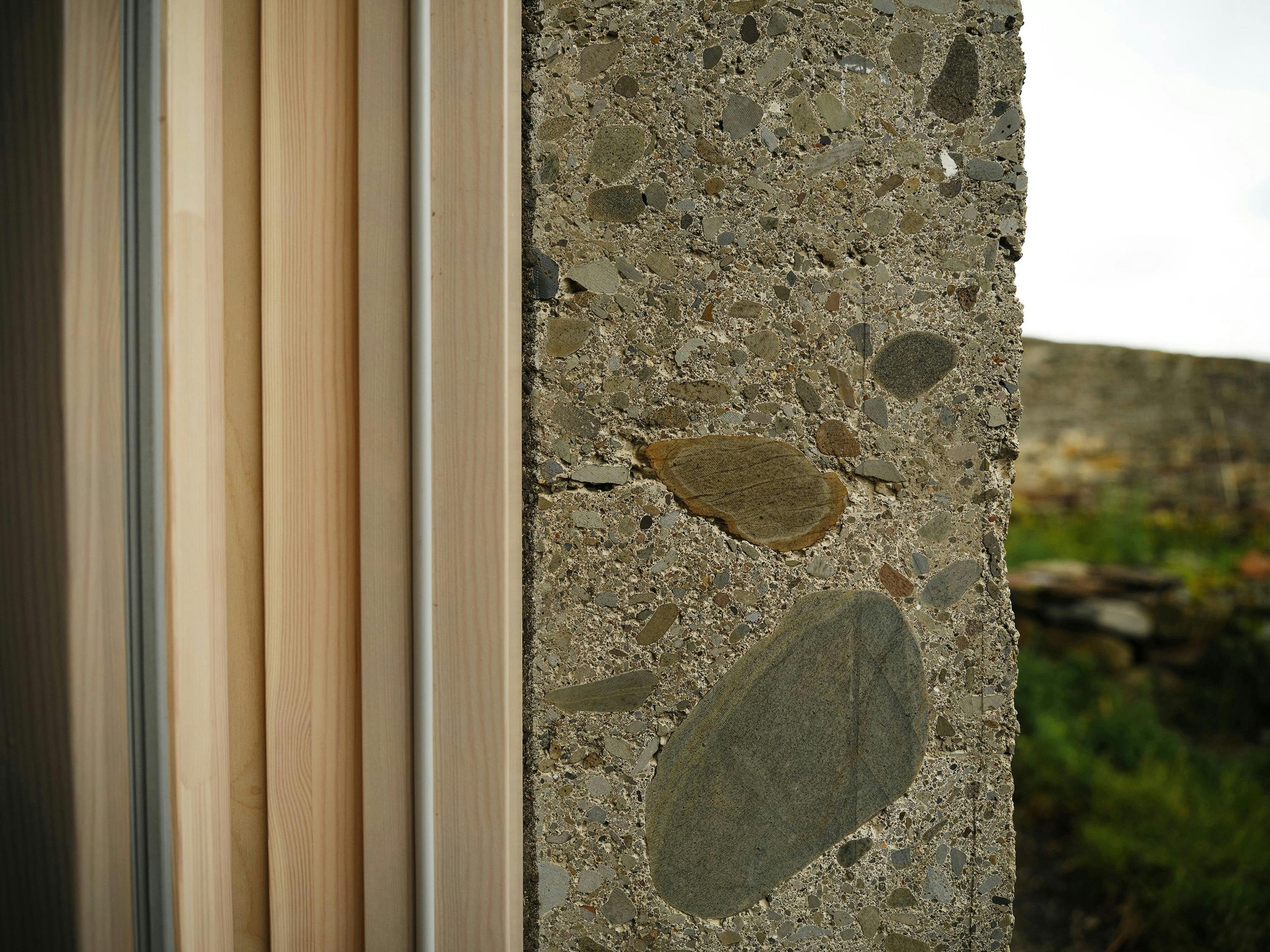 A close-up, straight-on photograph of a building detail, showing the junction between a wooden door frame and a concrete wall. The wooden door frame consists of multiple narrow, light-coloured wooden slats with a light grey trim. The concrete wall has a rough, textured surface with exposed aggregate, featuring various sizes and colours of pebbles and stones. A large, flat, grey stone is prominently visible in the centre of the concrete. The background shows a blurred outdoor scene with green grass and a rocky outcropping. The lighting is natural and highlights the textures of the wood and concrete.