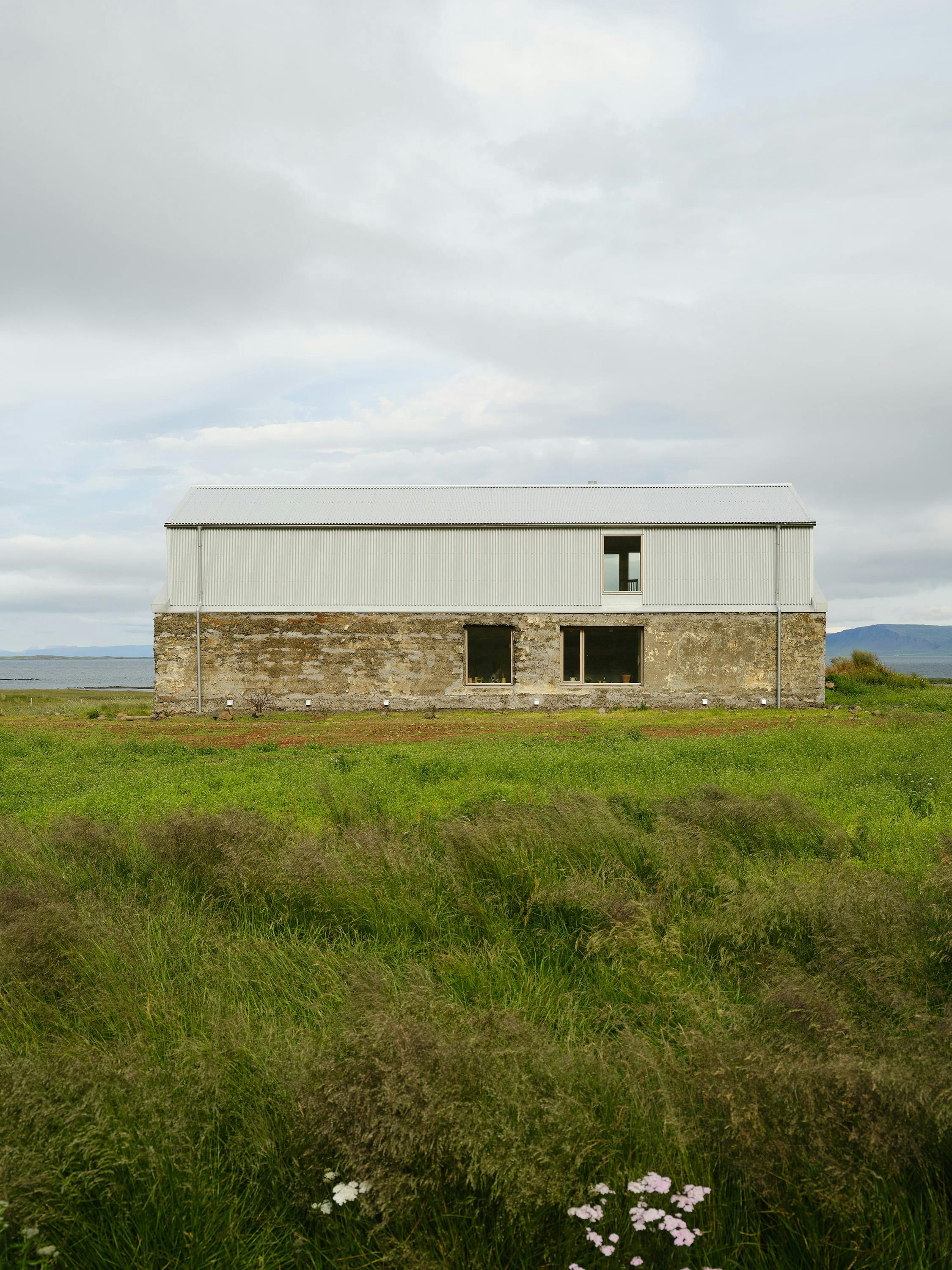 A straight-on, eye-level photograph of a building in a rural landscape. The building has a rectangular, barn-like shape with a light gray corrugated metal roof and upper walls. The lower walls are made of rough, textured stone. The building has a few small windows, including one with a distinctive 'T' shaped division. The building is situated in a field of tall, green grass with purple and white wildflowers in the foreground. A body of water and distant hills are visible in the background under a cloudy sky. The overall aesthetic is rustic and minimalist, showing the contrast between the building's modern materials and the natural landscape.