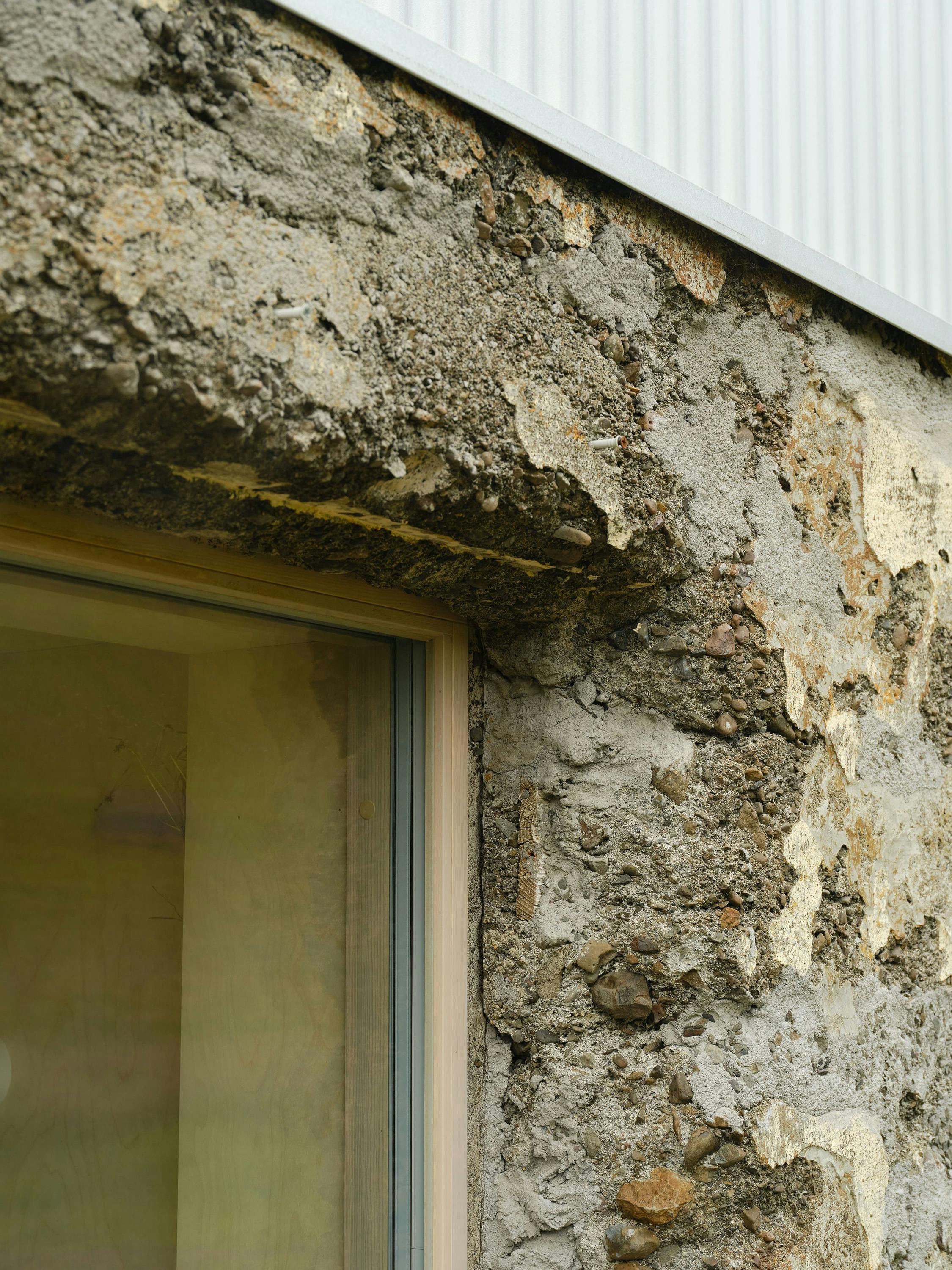 A close-up, slightly angled photograph of a building exterior detail. The image focuses on the junction between a rough, textured stone wall and a smooth metal flashing. The stone wall is grey and shows signs of weathering and erosion, with patches of lighter material and visible stones embedded in the mortar. A light-coloured window frame is partially visible at the bottom of the image. A smooth, light-coloured metal flashing runs along the top edge of the stone wall. The lighting is natural and highlights the textures and materials. The overall aesthetic is rustic and textural, showing the contrast between the natural stone and the manufactured metal
