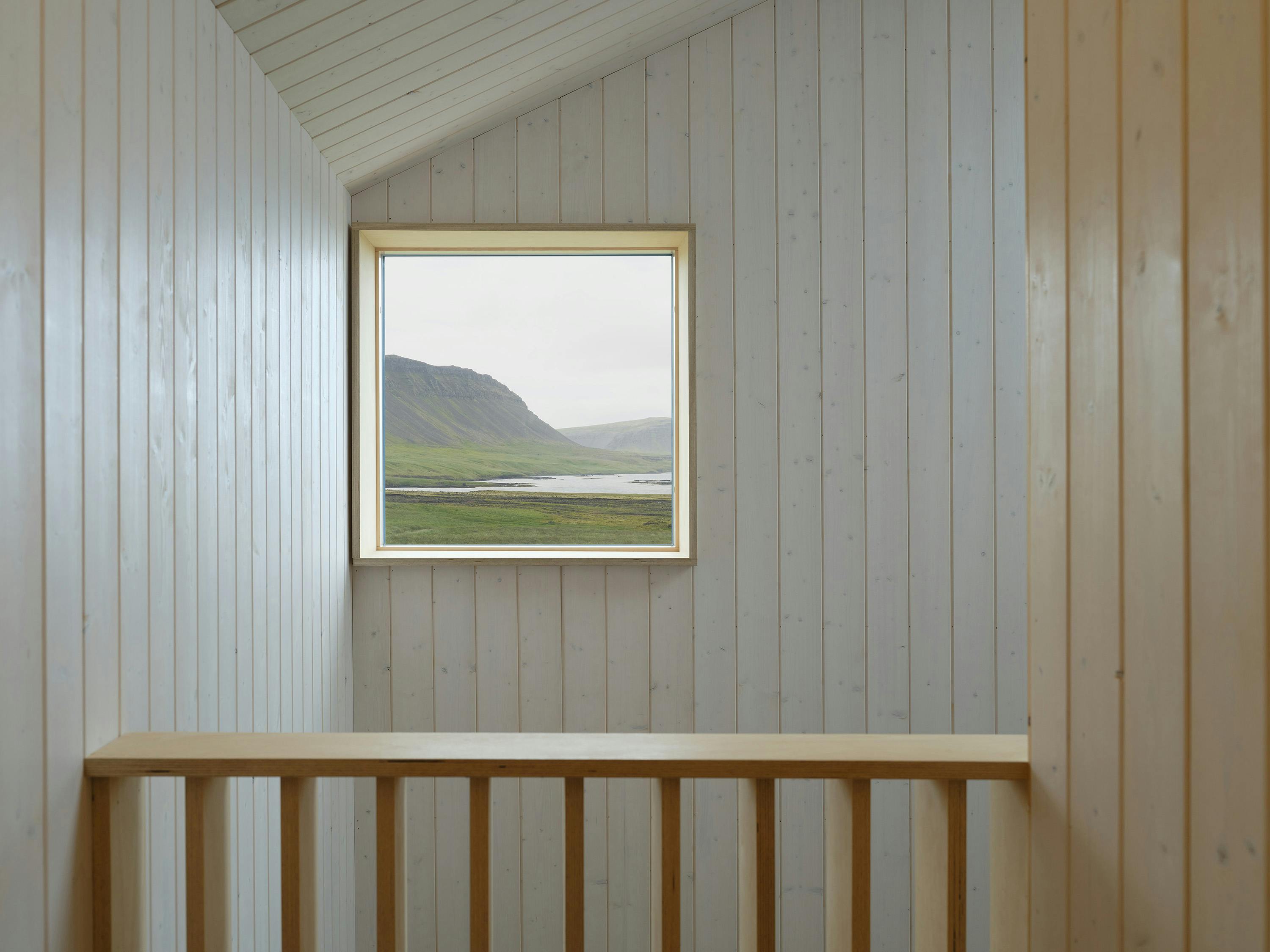 A slightly angled, eye-level photograph of an interior space with a view through a window. The space features light-colored, vertically paneled wood walls and a sloped ceiling. A rectangular window is centered on the far wall, offering a view of a green landscape with a body of water and distant hills. A light-colored wooden railing with vertical slats is visible in the foreground. The lighting is soft and natural, highlighting the textures of the wood and the view outside. The overall aesthetic is minimalist and natural, with a focus on simple materials and the surrounding landscape.