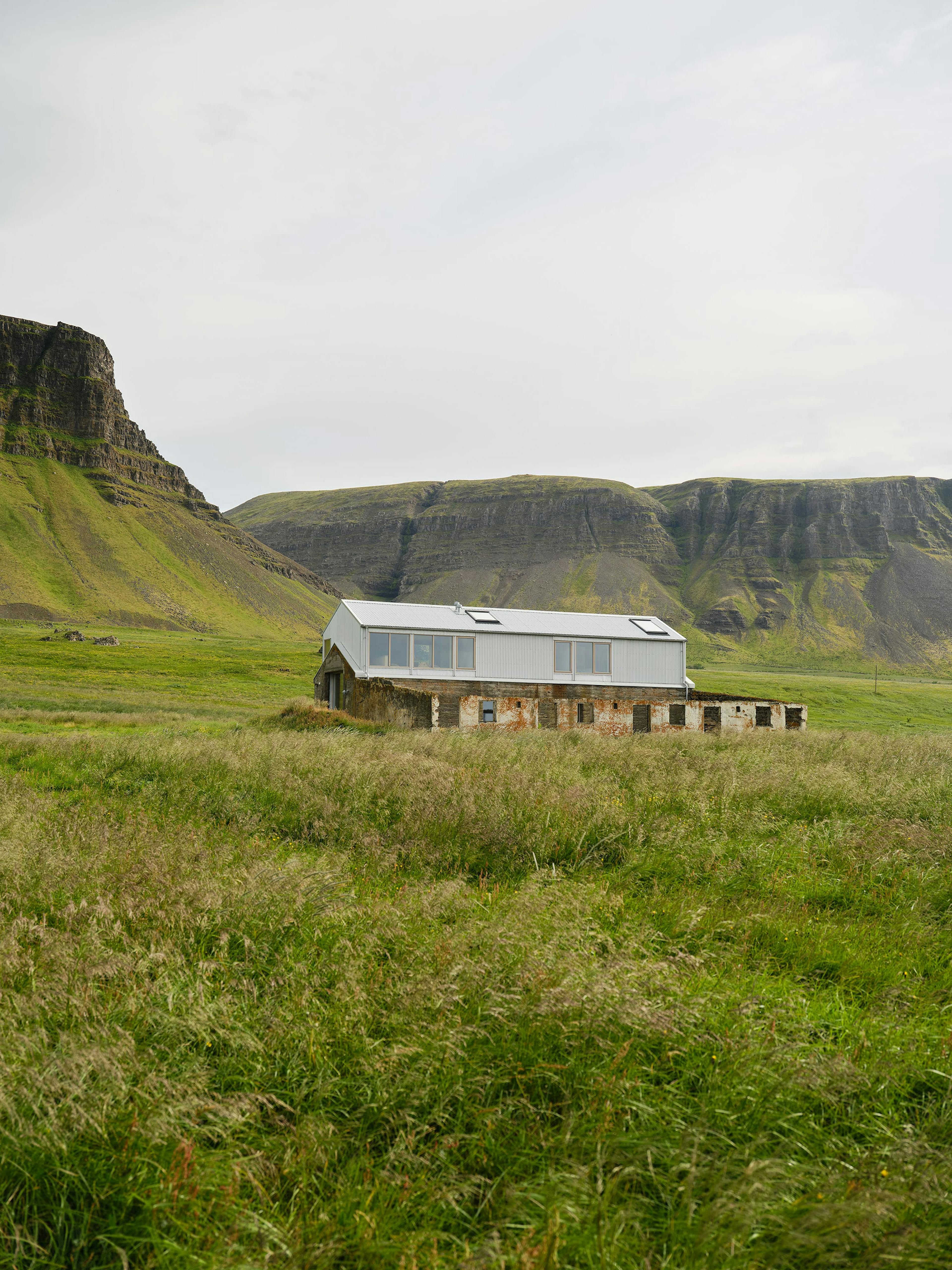 A wide, scenic shot of a building situated in a vast, grassy field. The building is a long, single-story structure with a metal roof and light-colored siding. It features several windows along its length. The building appears to be partially dilapidated, with some sections showing signs of age or damage, particularly at the base. The field is covered in tall, swaying grasses and wildflowers, creating a natural foreground. In the background, a large, rocky hill or mountain range rises, with a mix of green vegetation and exposed rock. The sky is overcast and gray, contributing to a sense of remoteness and tranquility. The image conveys a feeling of isolation and the integration of architecture into a natural, rugged landscape.