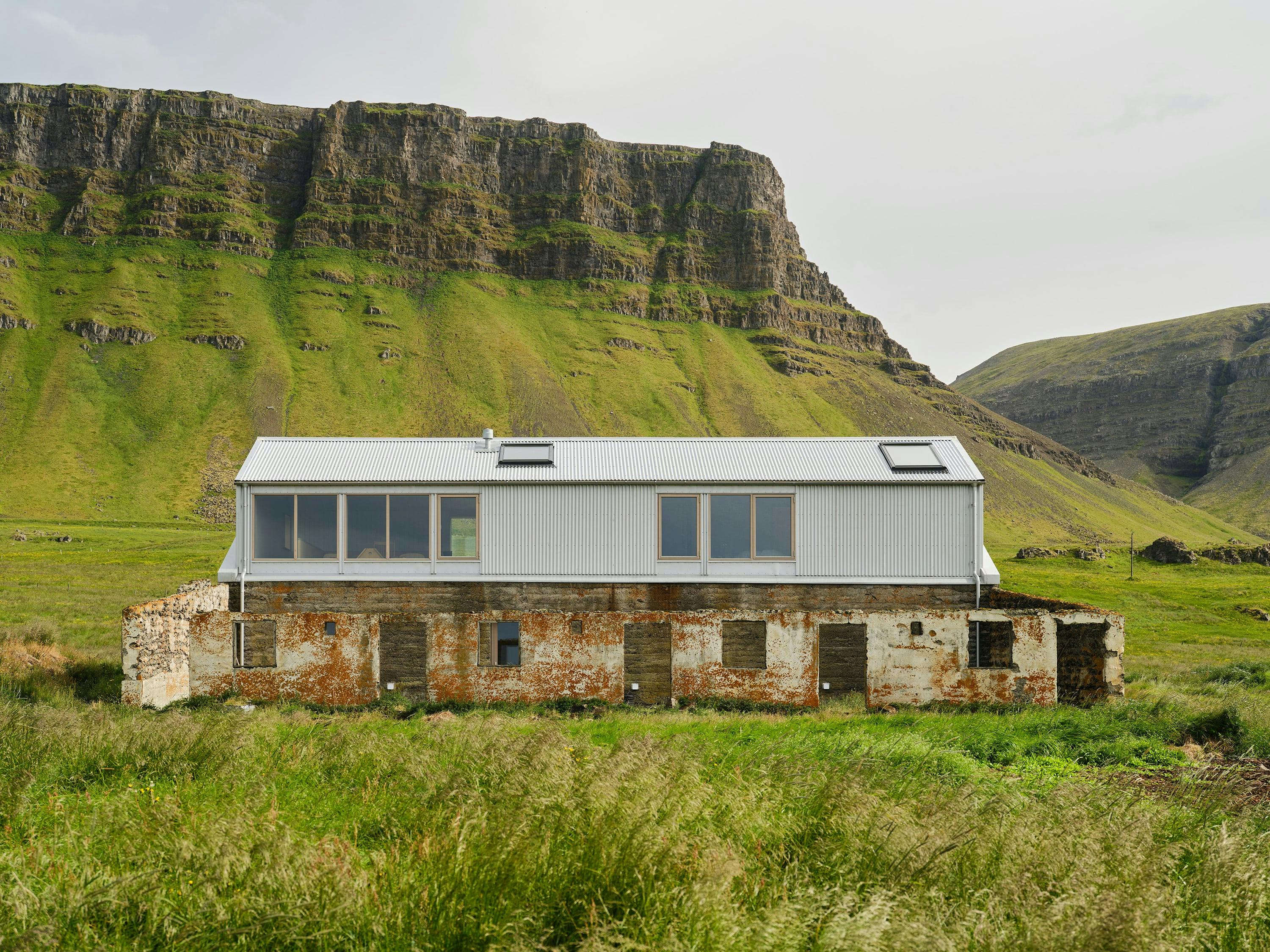 A straight-on shot of a two-story building set against a backdrop of a large, rocky cliff. The building features a striking contrast between its lower and upper sections. The lower section appears to be a weathered, stone structure with visible signs of age and deterioration, featuring multiple small, rectangular windows and doorways. The upper section is a modern addition with light gray or white corrugated metal siding, featuring larger windows and a metal roof. The building is situated in a grassy field with tall, wild grasses in the foreground. In the background, a large, rocky cliff with green vegetation dominates the scene, creating a dramatic backdrop. The sky is overcast and gray. The image conveys a sense of architectural juxtaposition, blending old and new styles in a natural, rugged setting.
