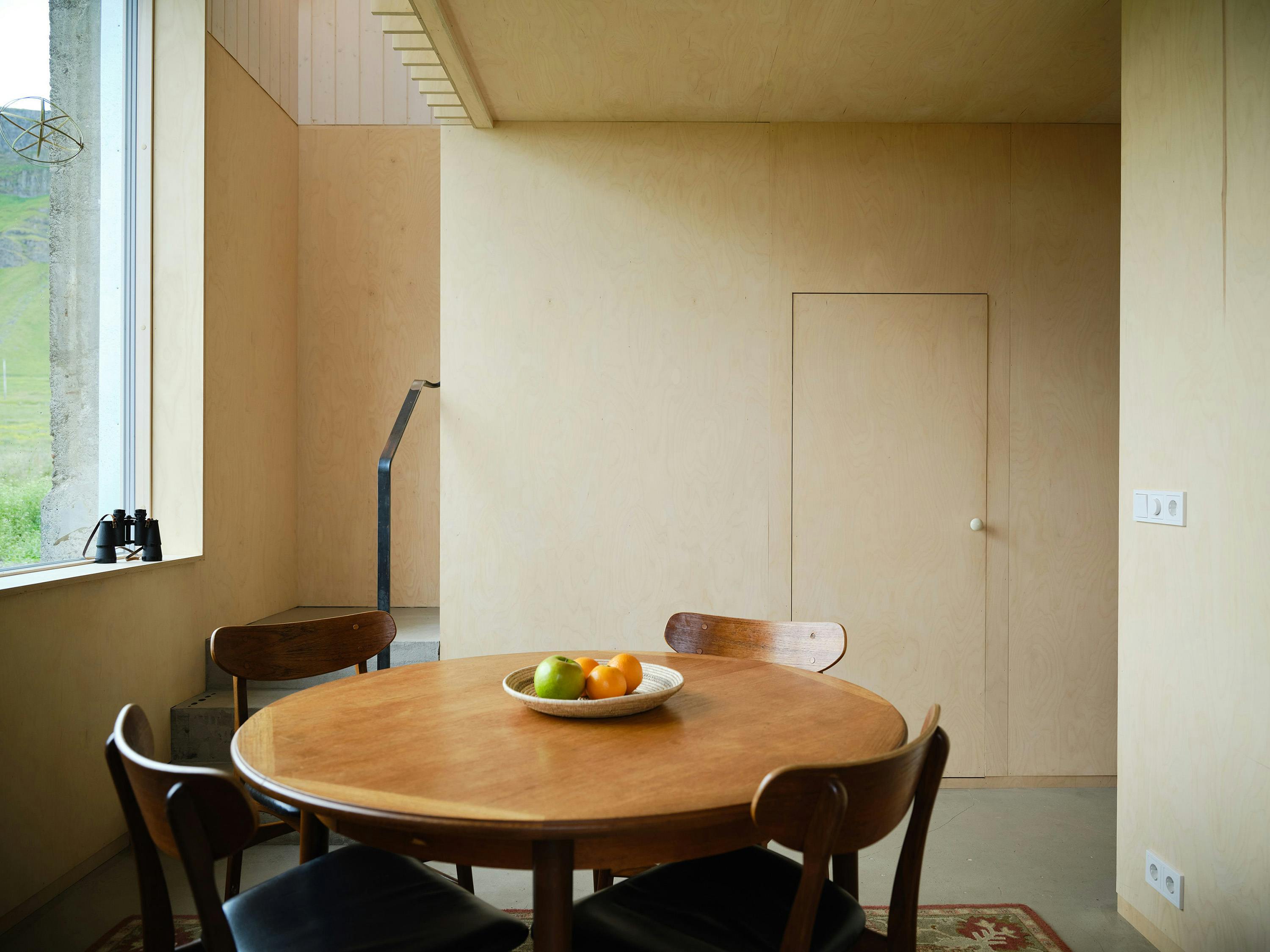 An interior shot of a minimalist dining area with a focus on natural materials. The room features light-colored plywood walls and ceiling, creating a warm and simple aesthetic. A round wooden table is centered in the space, with four chairs surrounding it. A white bowl containing oranges and a green apple sits on the table. In the background, a narrow staircase with a black metal railing is visible, leading to an upper level. A small, flush door is integrated into the wall on the right side of the image. A window on the left reveals an outdoor view of a green landscape and a cloudy sky. A pair of binoculars is placed on the windowsill. The floor appears to be concrete or a similar material. A small rug with a patterned design is partially visible in the bottom right corner. The lighting is soft and diffused, highlighting the natural textures of the wood. The overall scene conveys a sense of Scandinavian-inspired minimalism and a connection with nature.