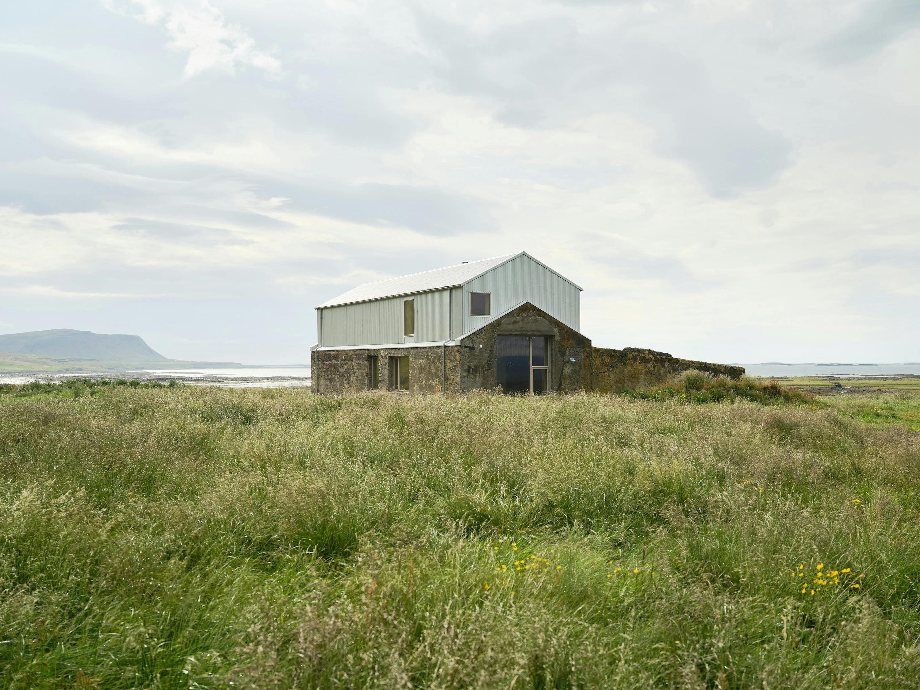 A renovated barn or farm building with a modern metal addition sits in a field of tall grasses in West Iceland. The building features a concrete base and a light-colored metal upper level with a pitched roof. A large glass door is visible. The sky is cloudy and a body of water and distant hills are in the background.