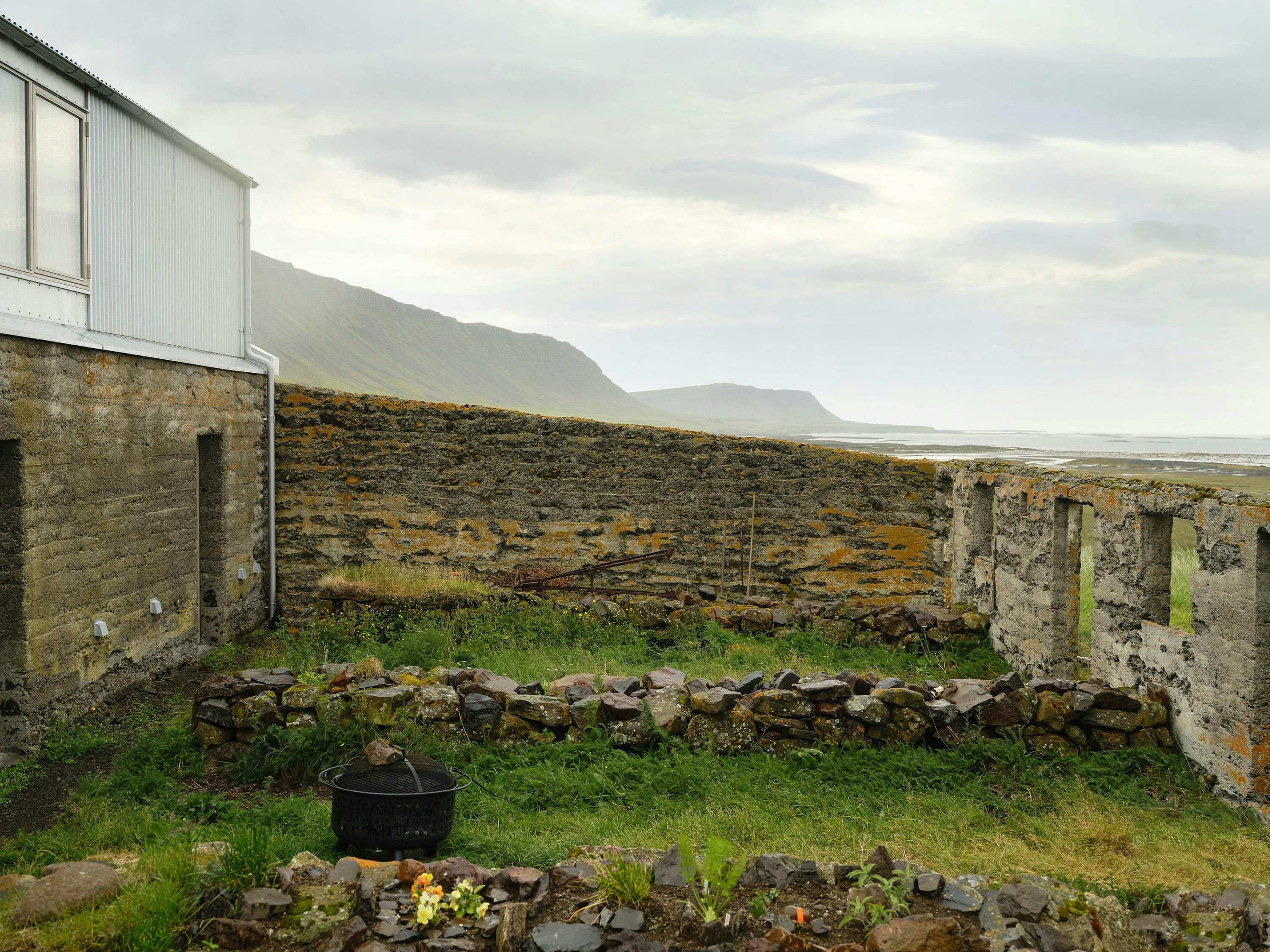 A slightly angled shot of a building with a mix of old and new architecture, set against a backdrop of a coastal landscape. The building features a modern section on the left with light-colored metal siding and a sloped roof. Adjoining this section is an older, weathered stone structure with visible signs of age and deterioration, including missing sections of wall and exposed brickwork. The stone structure appears to be a ruin or a partially preserved building. In the foreground, a small courtyard area is defined by low stone walls, with patches of green grass and moss growing between the stones. A black metal bucket sits in the courtyard, and some flowers are visible near the base of the stone wall. In the background, a rocky hill or mountain range rises, leading down to a coastal area with a body of water extending to the horizon. The sky is overcast and gray. The image conveys a sense of history and the integration of modern architecture with the remnants of older structures in a natural, outdoor setting.