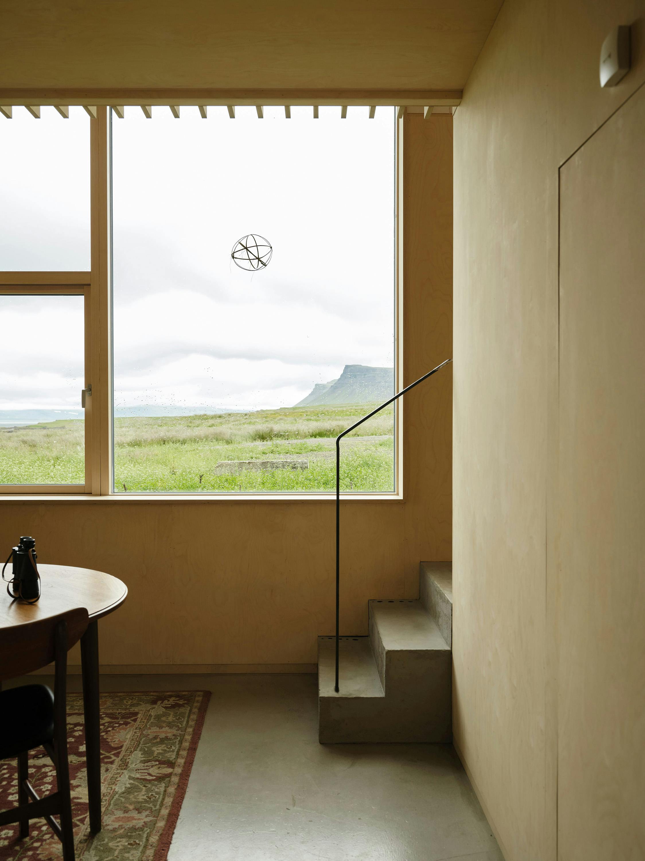 An interior shot of a room with a minimalist design and a large window overlooking a landscape. The room features light-colored plywood walls and ceiling, creating a warm and simple aesthetic. A large rectangular window dominates the far wall, offering a panoramic view of a green field leading to a body of water and a distant mountain range under a cloudy sky. A spherical metal sculpture hangs in the center of the window. On the left side of the room, a round wooden table with a dark chair is partially visible, along with a patterned rug on the floor. A black metal teapot sits on the table. On the right side, a set of concrete steps leads upwards, with a black metal handrail. A flush door is integrated into the wall on the right. A small, white rectangular object is affixed to the upper right wall. The lighting is soft and diffused, highlighting the natural textures of the wood and the concrete. The overall scene conveys a sense of Scandinavian-inspired minimalism and a connection with nature