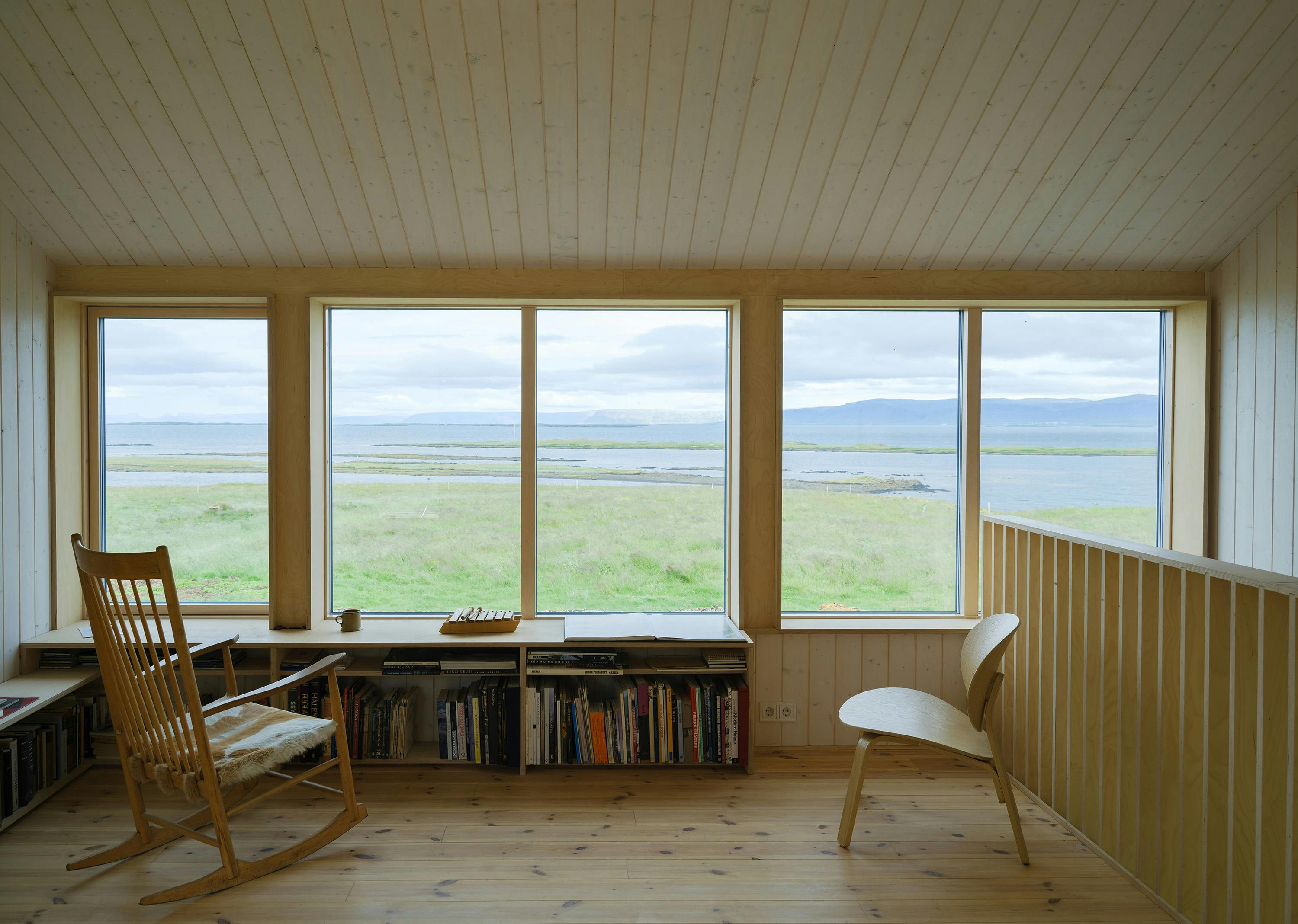 An interior shot of a bright, minimalist room with a panoramic view of a landscape. The room features light-colored wooden plank walls and ceiling, creating a warm and natural atmosphere. Three large windows dominate the far wall, offering a view of a grassy field leading to a body of water, possibly a lake or ocean, with a distant shoreline and a cloudy sky. The room has light-colored wooden flooring. On the left side of the room, a wooden rocking chair faces the windows, positioned next to a built-in bookshelf filled with books and objects. Another wooden chair with a curved back is on the right side of the room, near a wooden railing. The railing suggests a slightly elevated platform or a change in floor level. The overall scene conveys a sense of tranquility and connection with nature, with a focus on simple, natural materials and a serene view.
