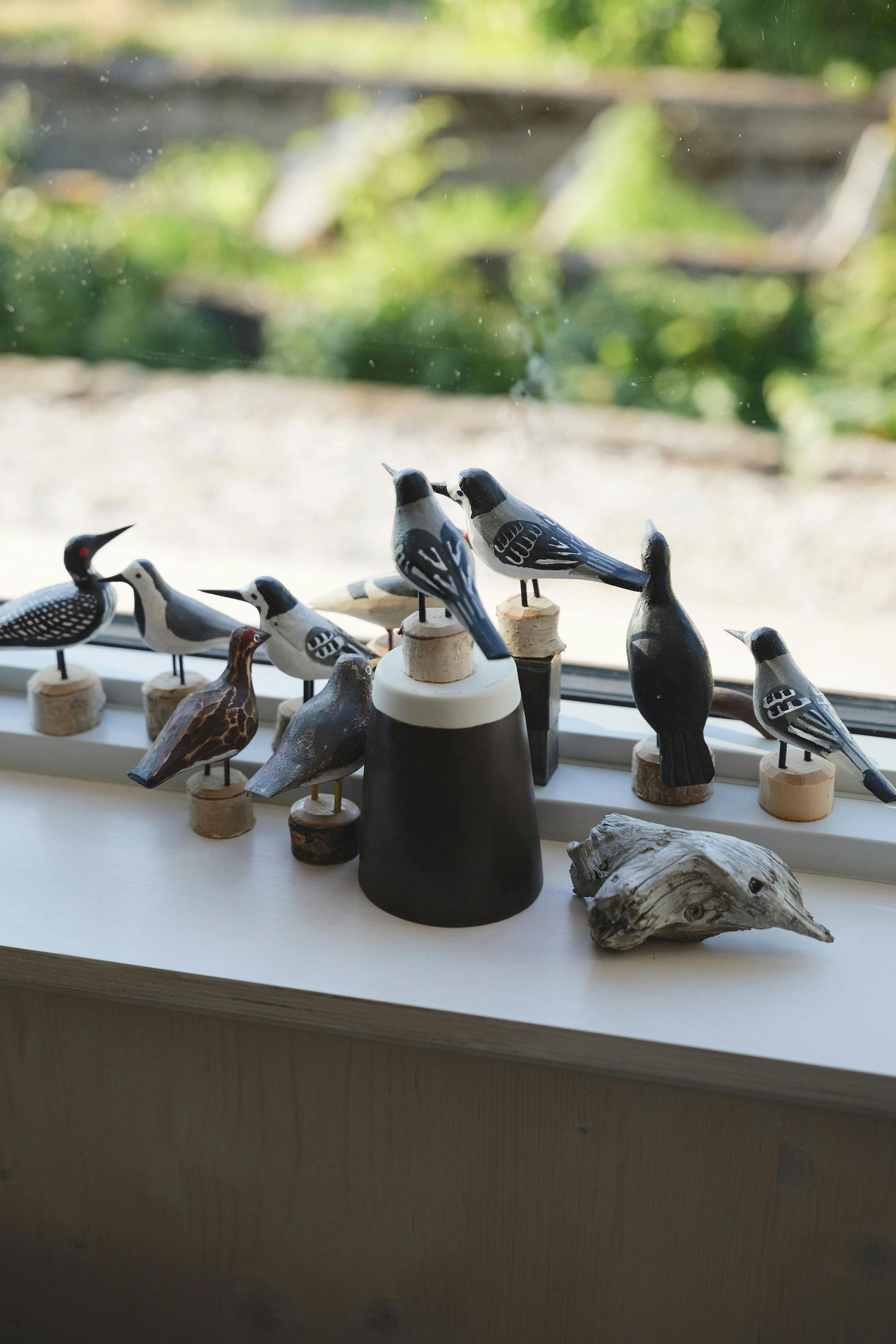 Nine carved wooden bird figurines of various sizes and colors are arranged on a white windowsill. A dark cylindrical object with a light-colored top and a piece of driftwood are also present. A blurred green landscape is visible through the window.