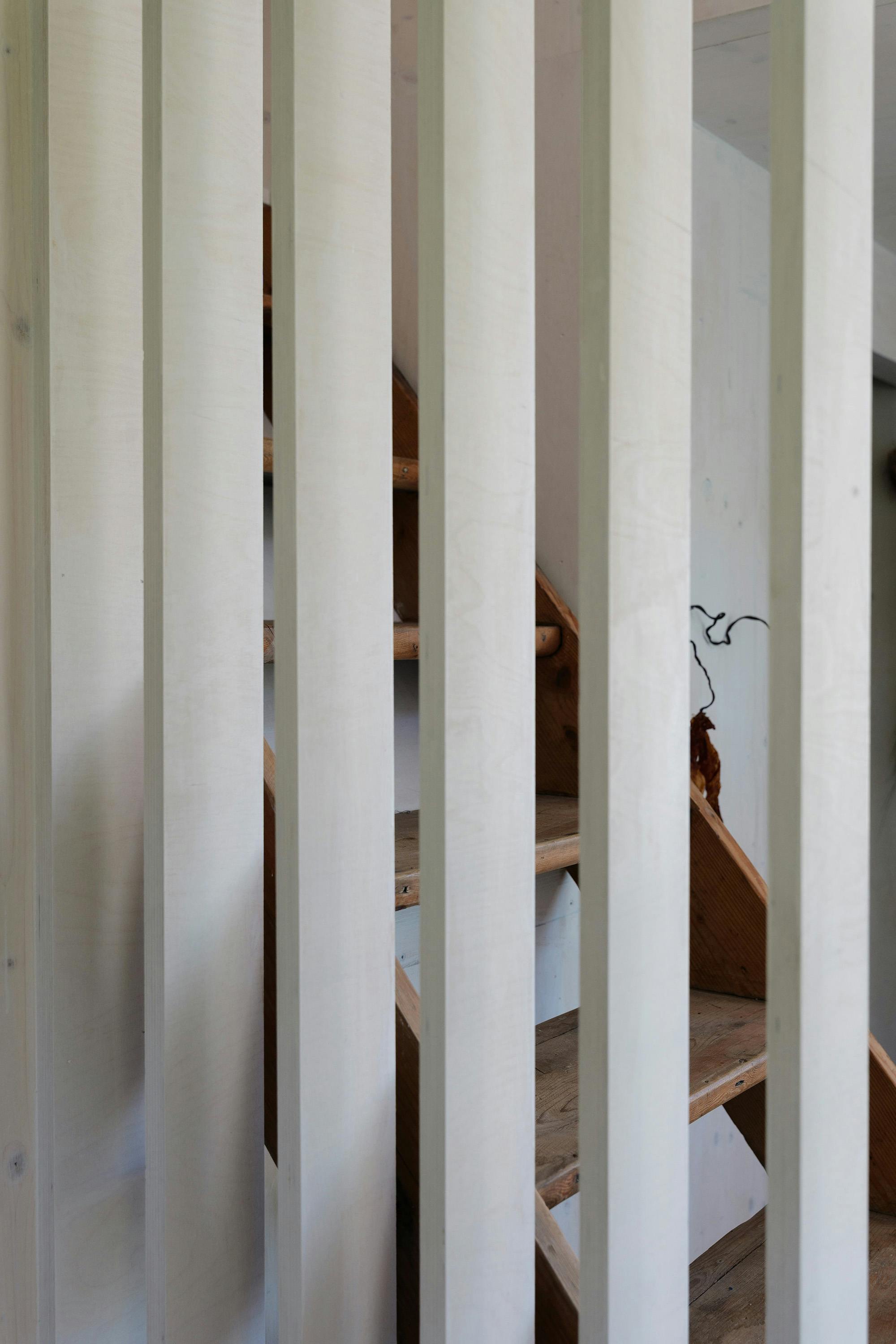 A close-up shot of a slatted wooden partition with light-colored slats. A wooden staircase is visible through the gaps between the slats, leading to an unseen area. The background is out of focus.