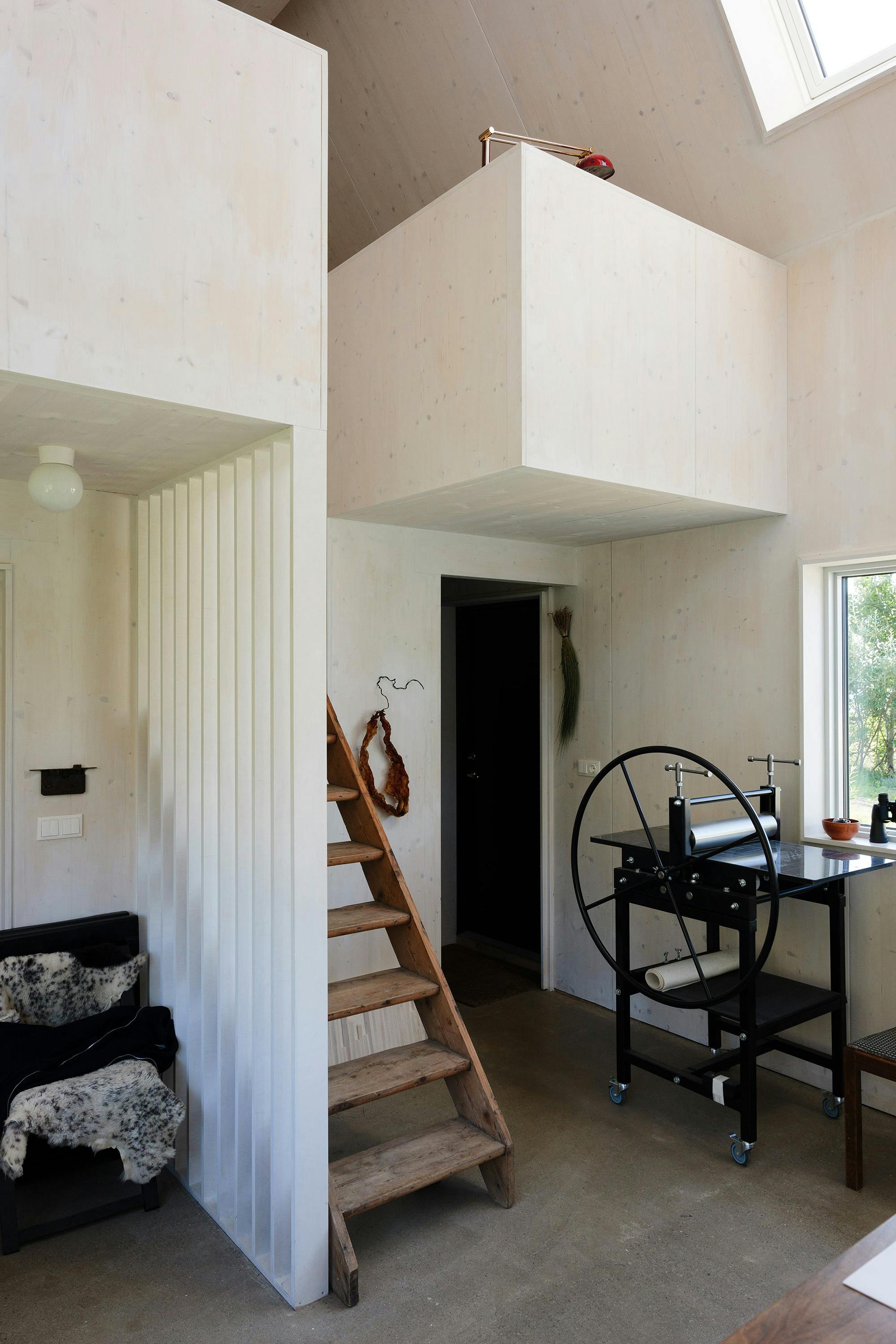 An interior room with light-colored walls and a concrete floor features a wooden staircase leading to a loft space. A black printing press sits on a table with wheels. A black door is visible, and a decorative object hangs on the wall. A black chair with a patterned throw is on the left.