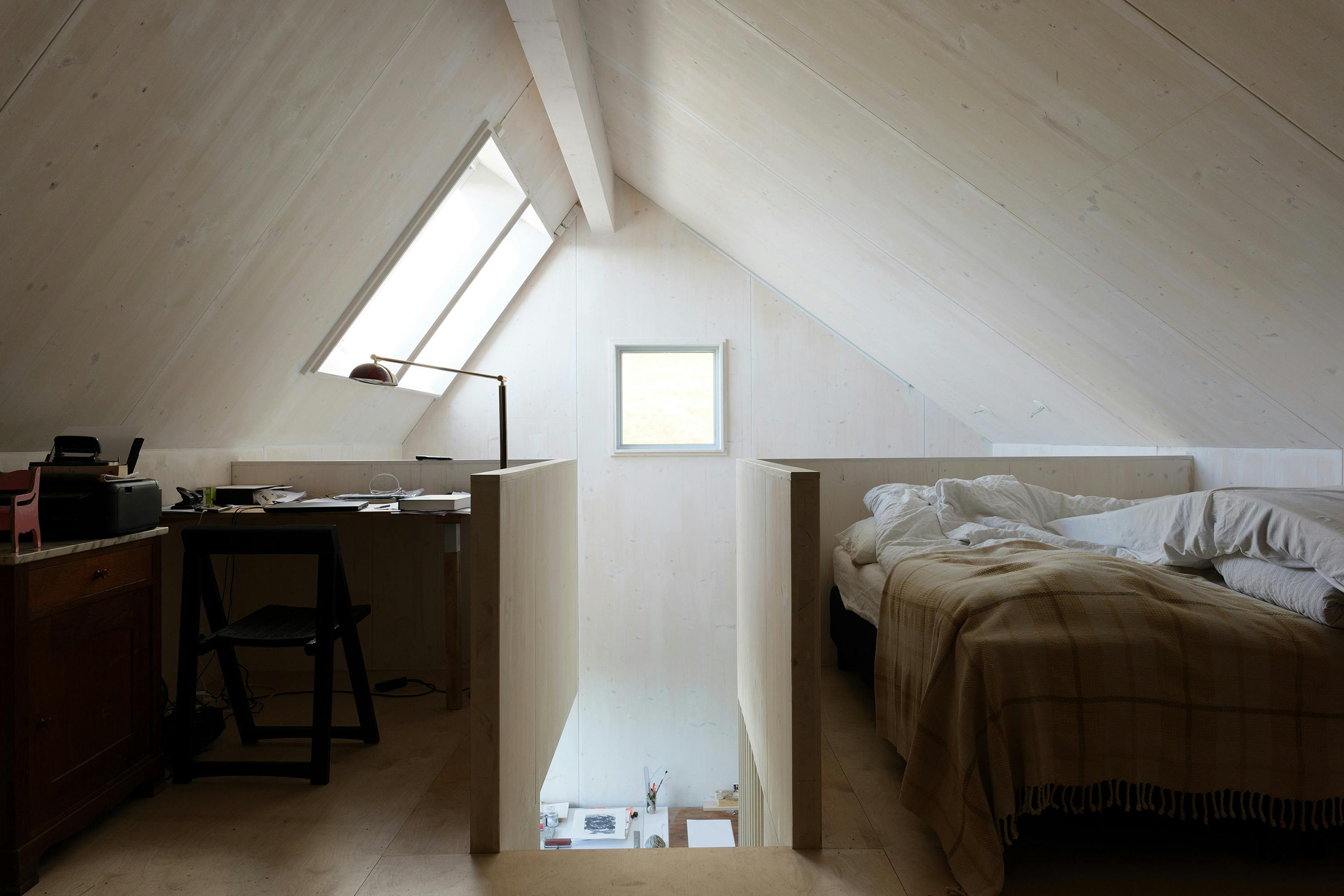 A room with sloped ceilings and light-colored wood paneling features a workspace with a desk and chair on one side, and a bed with a brown blanket on the other. A skylight and a window provide natural light. A partial wall separates the two areas.