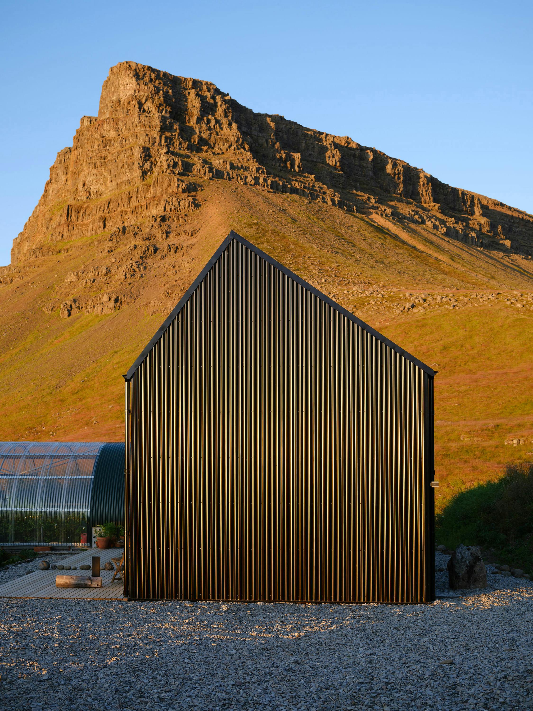 A modern black cabin with vertical ribbed siding and a pitched roof stands in a gravel clearing with a large, rocky mountain in the background. A greenhouse with a metal frame and translucent panels is partially visible to the left. The scene is bathed in warm, late-day sunlight. A project by London-based Studio Bua.