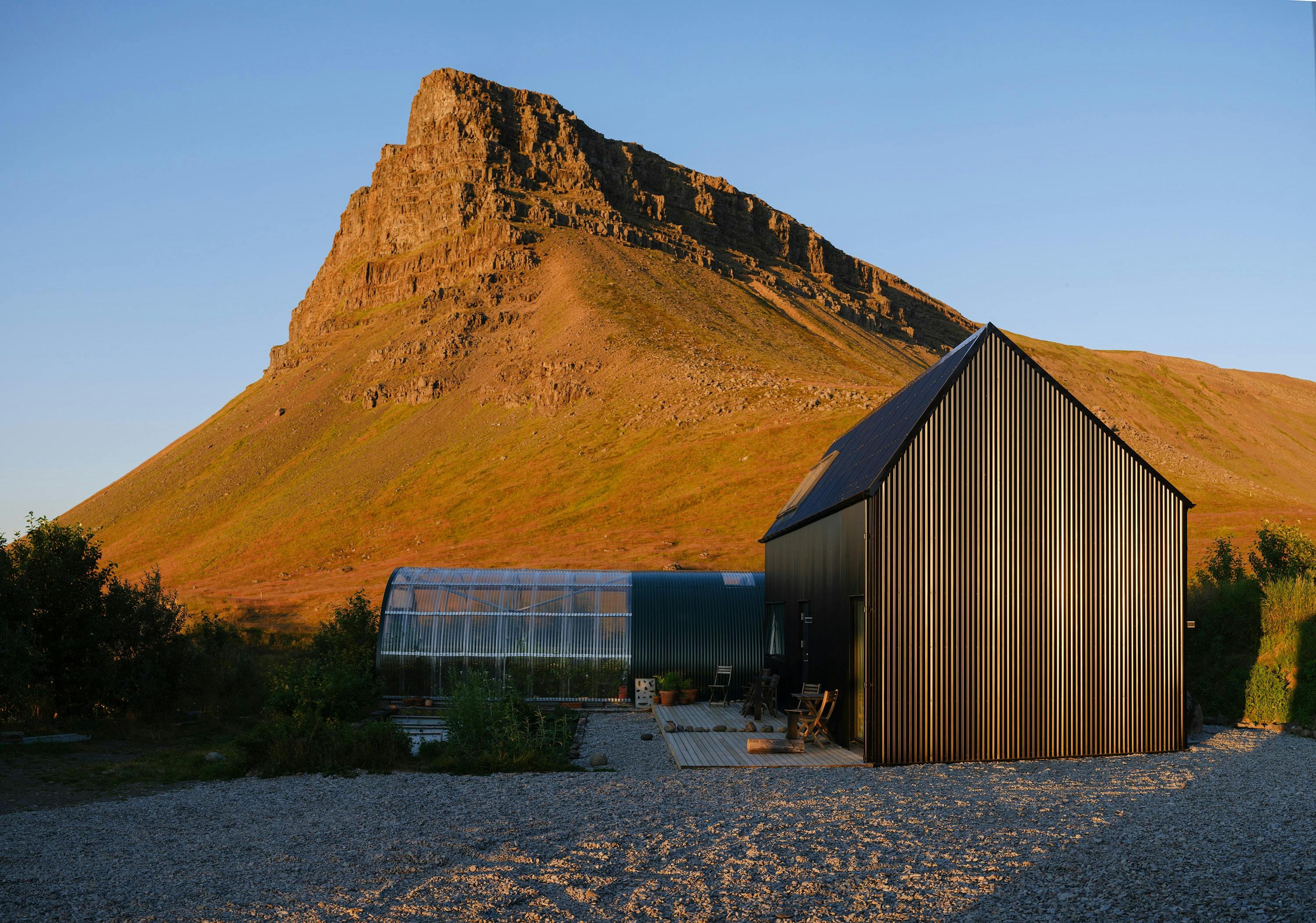 A modern black cabin with vertical ribbed siding stands in a gravel clearing with a large, rocky mountain in the background. A greenhouse is partially visible behind the cabin. The scene is lit by warm, late-day sunlight.