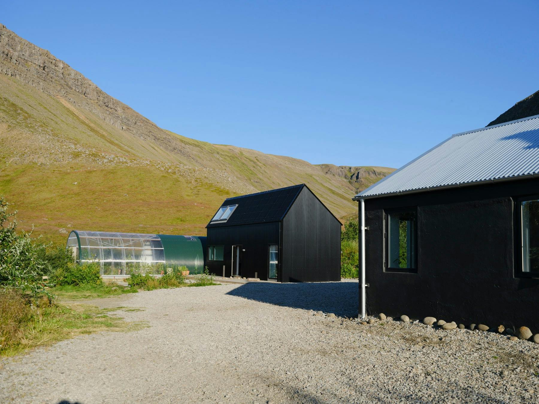 A gravel driveway leads to two modern black buildings with pitched roofs, including Guesthouse Nýp. A greenhouse with a metal frame and translucent panels is visible to the left. A mountain valley with rocky slopes is visible in the background. The sky is clear and blue.