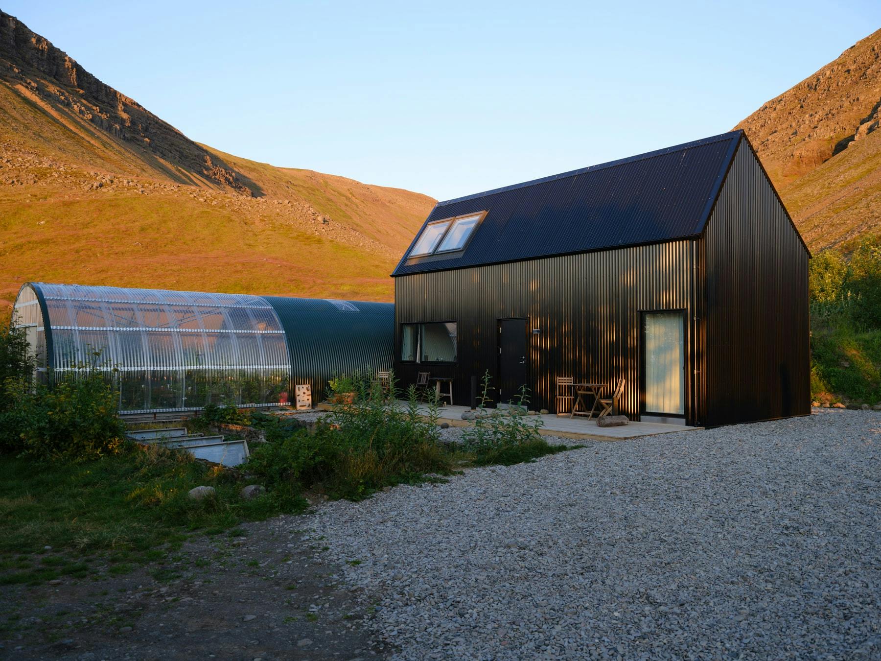 A modern black cabin with a pitched roof and ribbed siding stands in a gravel clearing with a large greenhouse to the left. The cabin features a small window on the upper level and a door and window on the lower level. A mountain valley is visible in the background. A project by London-based Studio Bua.
