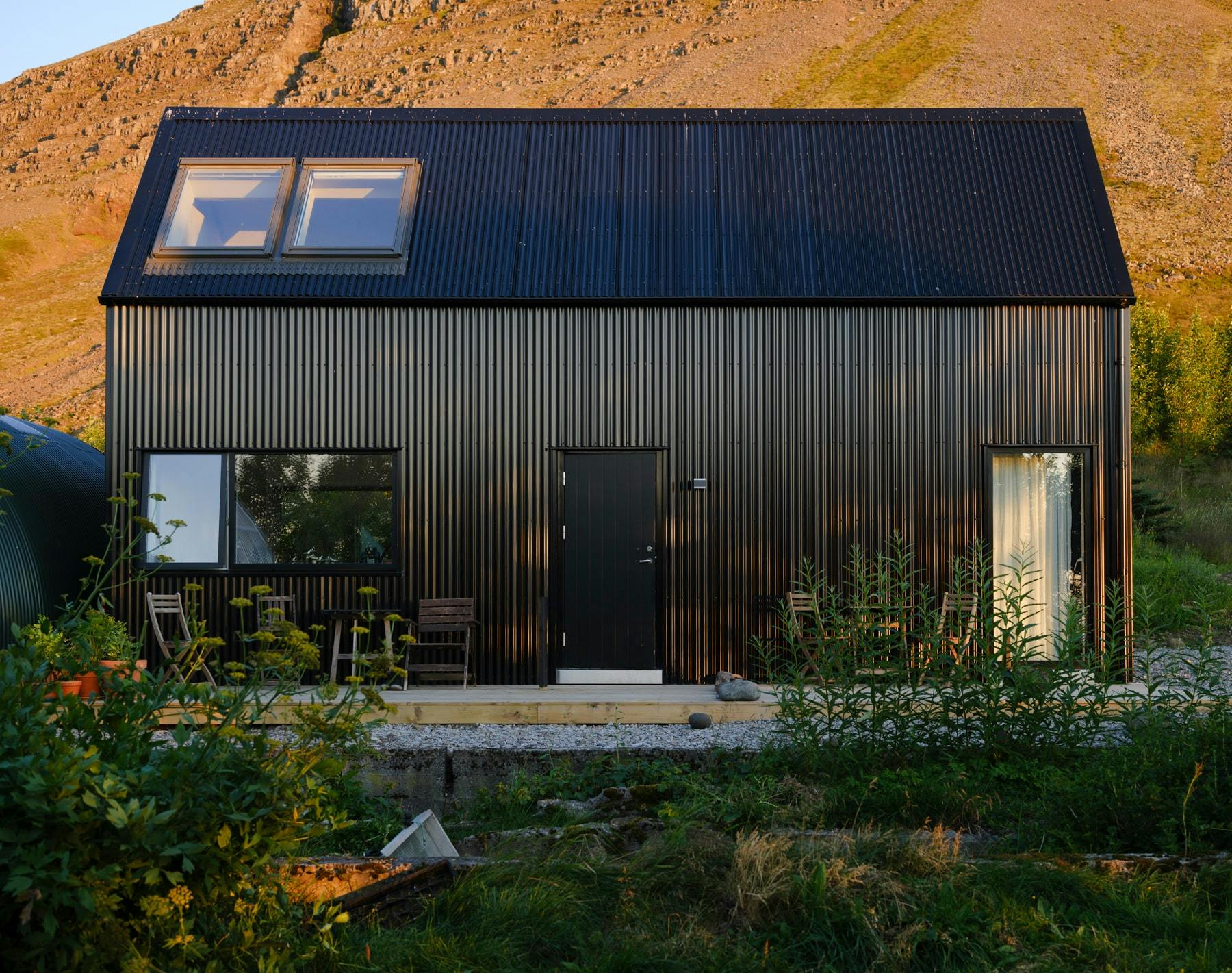 A modern black cabin with vertical ribbed siding stands in a gravel clearing with a large, rocky mountain in the background. A greenhouse is partially visible behind the cabin. The scene is lit by warm, late-day sunlight.