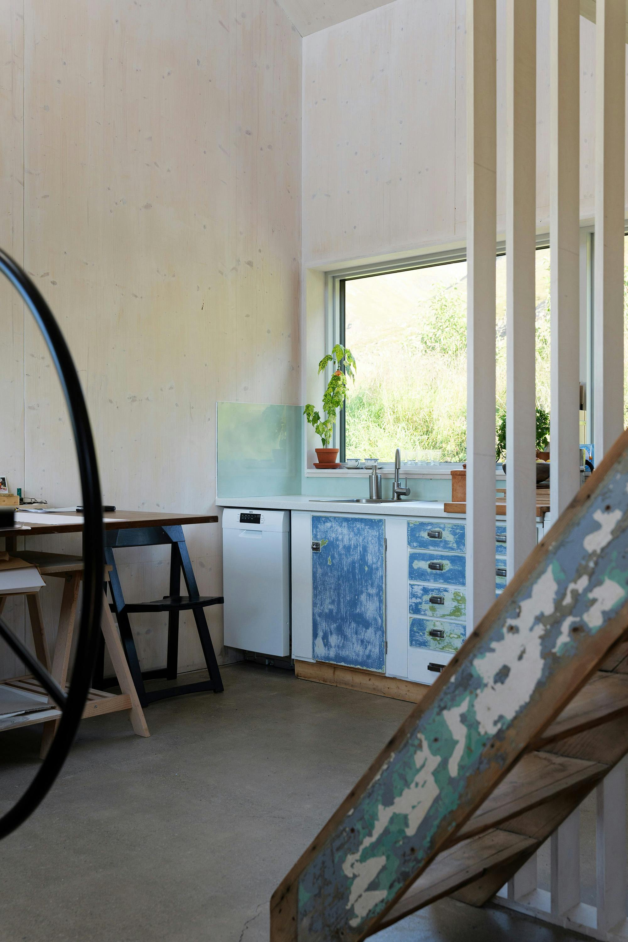 An interior room features a small kitchen area with white and distressed blue cabinets, a sink, and a mini-fridge. A window overlooks a green outdoor area. A wooden staircase with distressed paint is visible in the foreground, along with a black metal wheel and a desk. The room has light-colored wood paneling and a concrete floor.