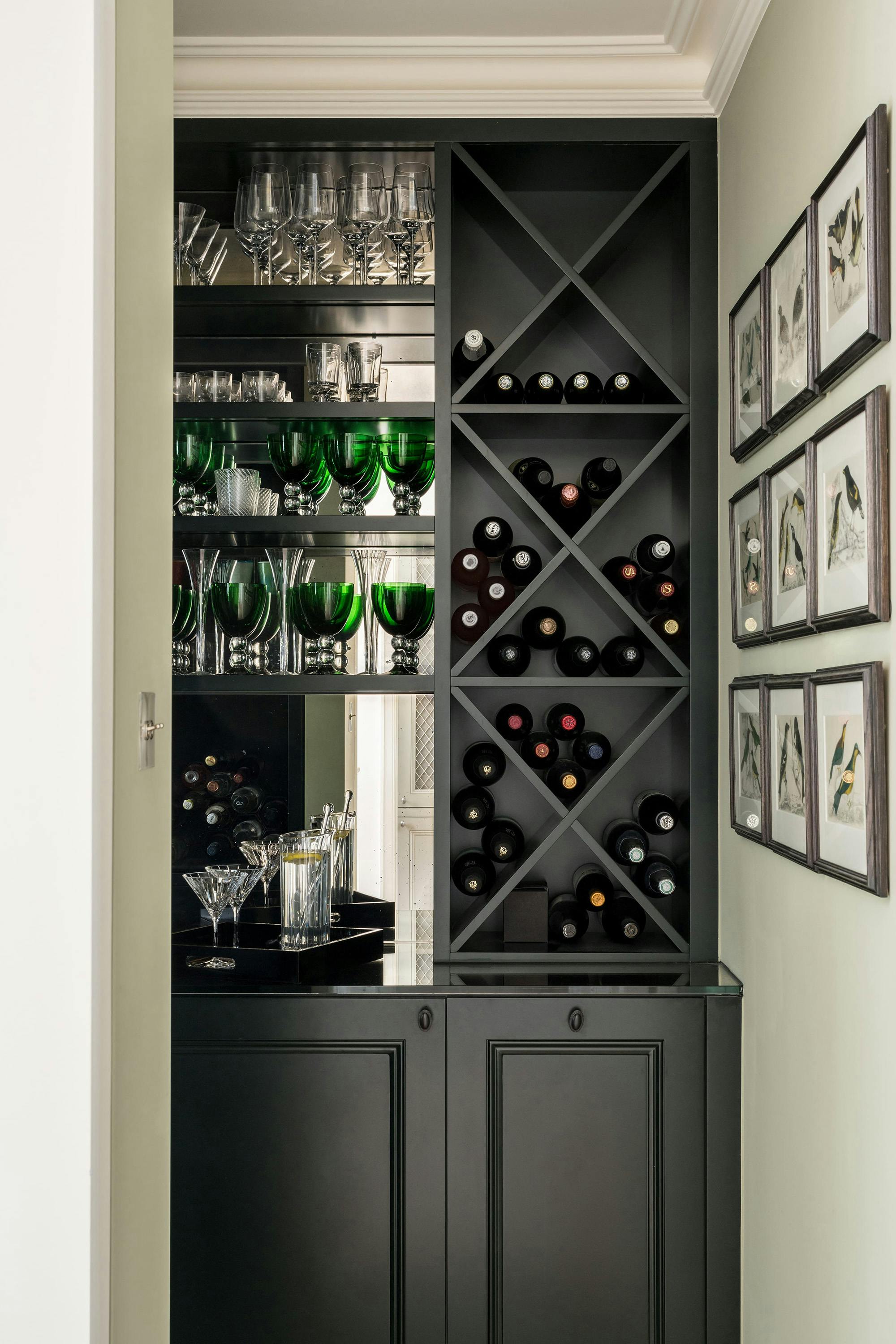 A dark, built-in bar cabinet features a glass-fronted upper section with shelves for glassware, including green glasses, and a wine rack with bottles. Framed artwork is displayed on the adjacent wall. The cabinet has closed doors at the bottom.