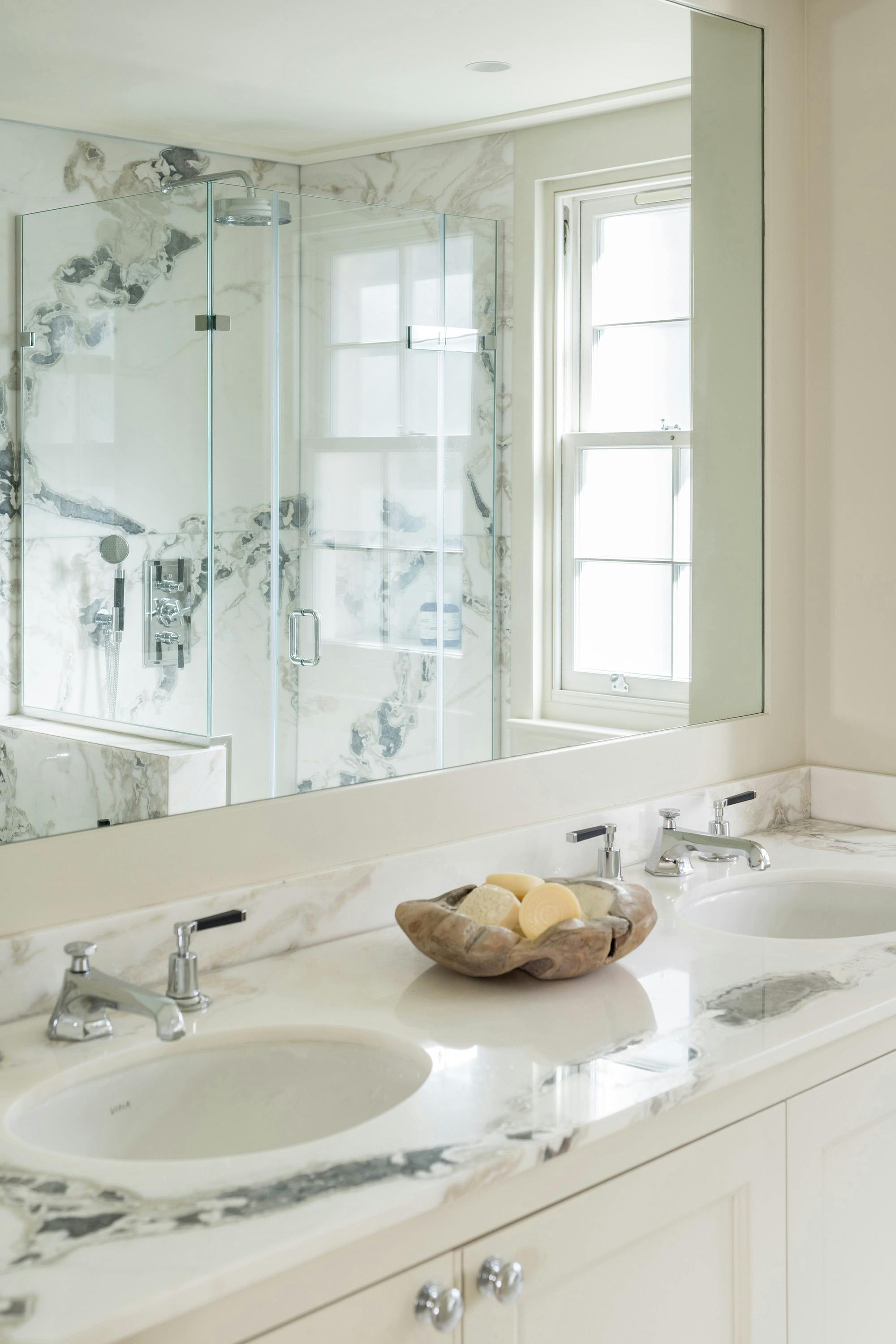 A bathroom with light-colored walls and a marble countertop with two sinks. A large mirror reflects a glass-enclosed shower with patterned marble walls and a window. A decorative bowl with soaps sits on the countertop.