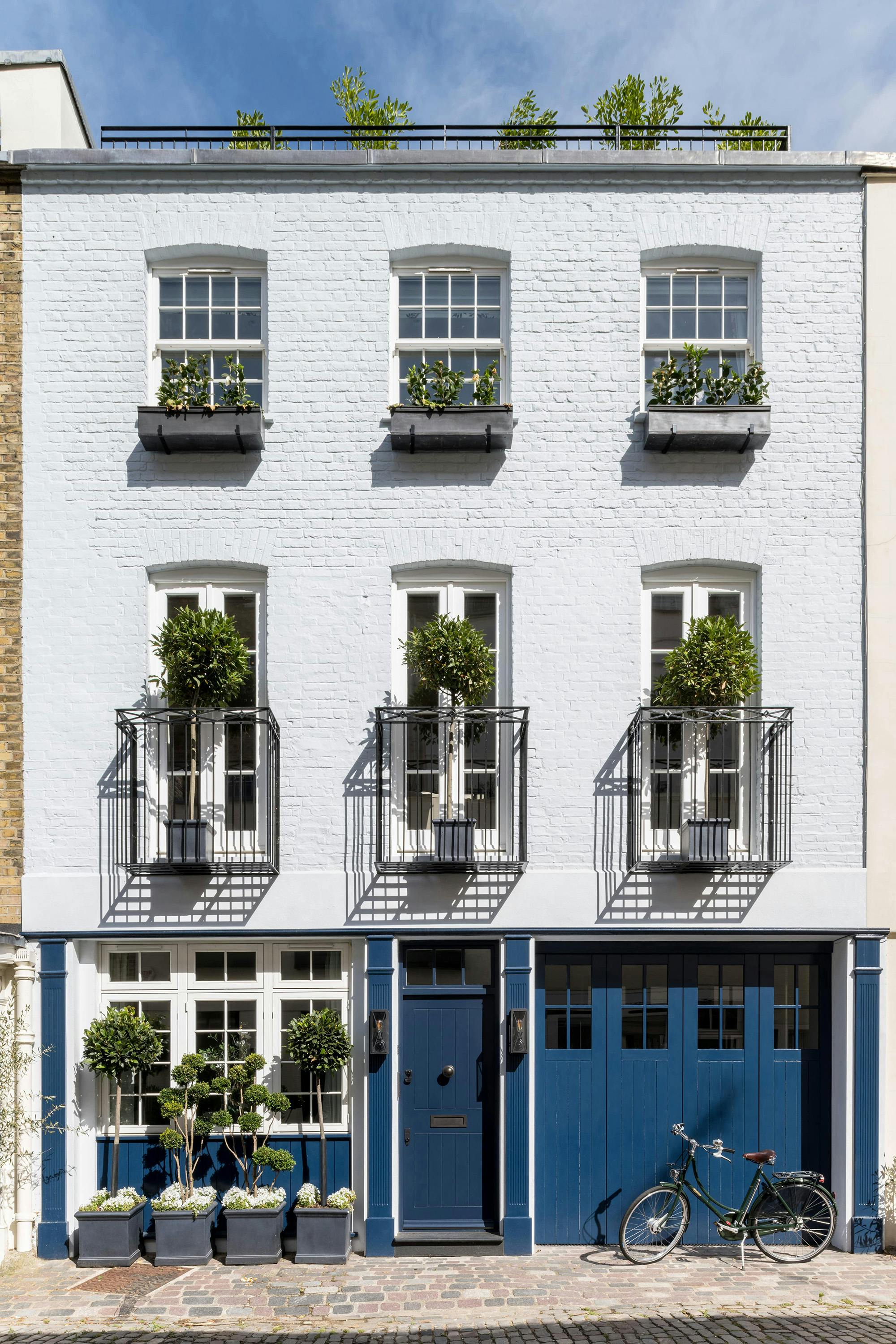 A three-story London townhouse with a white facade features traditional sash windows with flower boxes and wrought-iron balconies with potted trees. The ground floor has blue painted doors and garage doors, with potted plants and a bicycle parked on the cobblestone street.