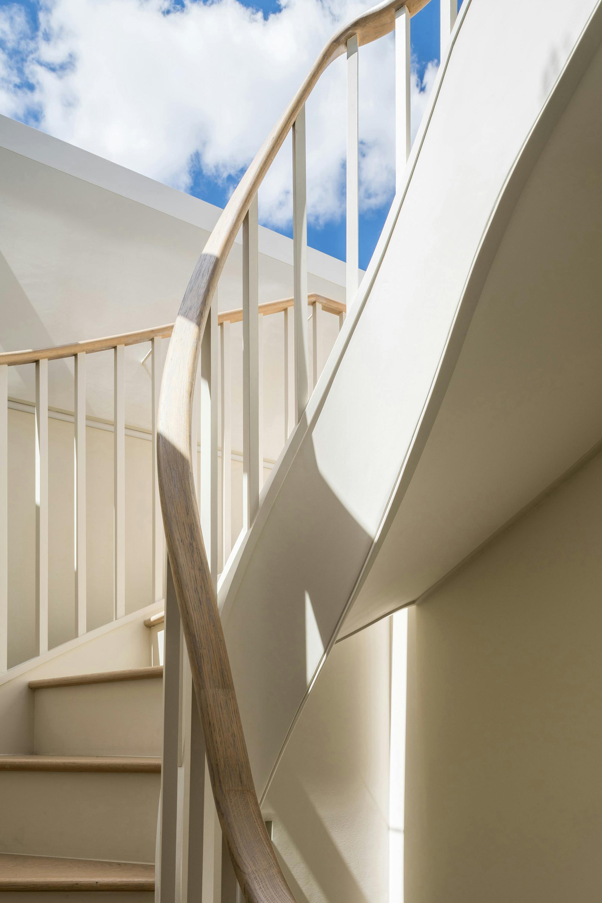 A detailed view of a spiral staircase with pale-colored steps and a light wooden handrail. The staircase curves upwards, and a section of white wall is visible. A bright blue sky with fluffy white clouds is visible in the upper part of the image, suggesting an outdoor or skylight view.