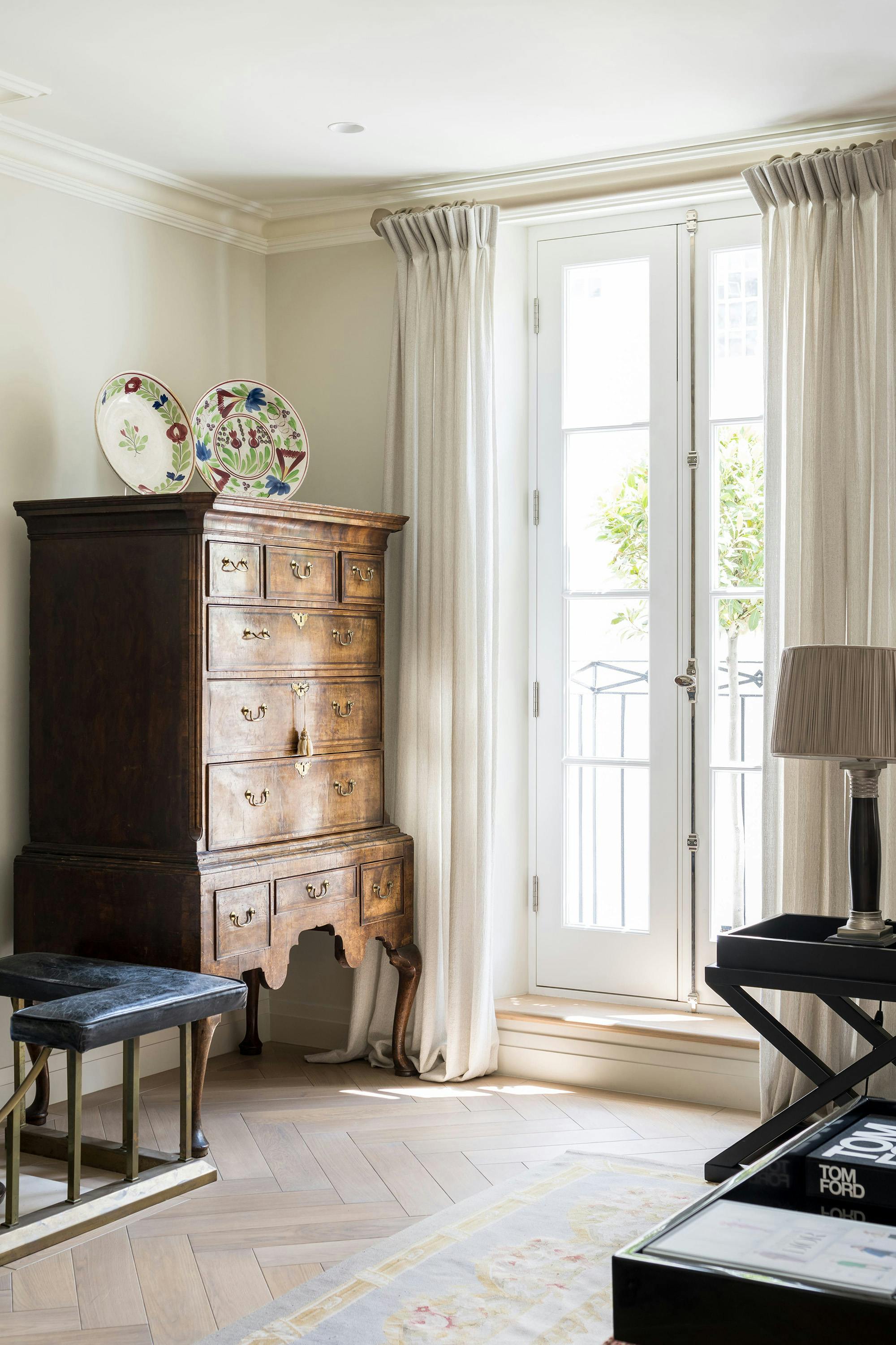 A room with light-colored walls and herringbone wood flooring features a wooden antique chest with drawers and decorative plates above. White French doors with sheer curtains provide natural light. A small stool with a blue cushion and a side table with a lamp are present.