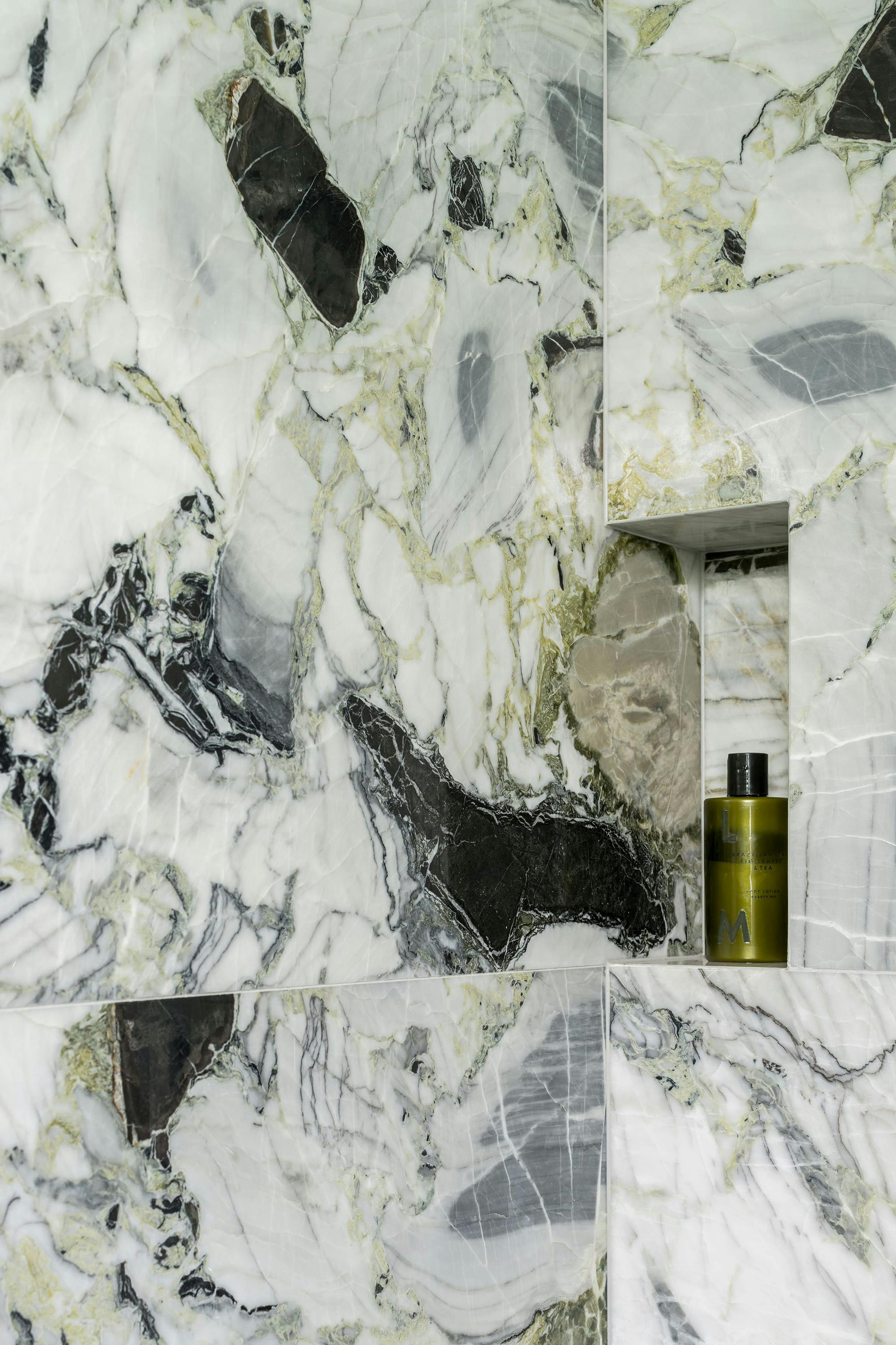 A detailed view of marble bathroom tiles with a swirling pattern of white, black, and grey. A small, recessed shelf in the wall holds a dark green bottle of liquid.