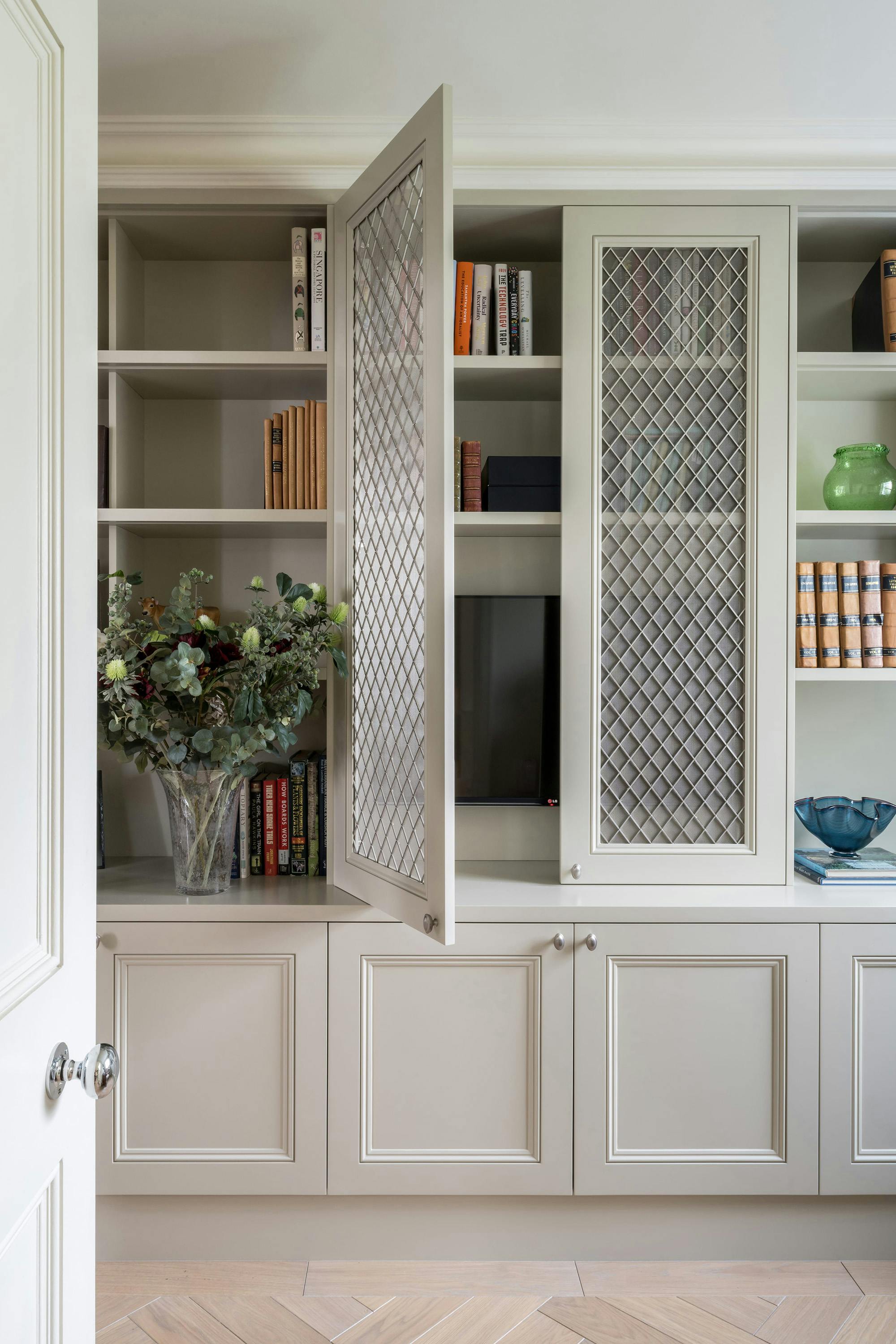 A built-in bookcase with light-colored shelves and wire mesh doors is featured in a room with herringbone wood flooring. The bookcase includes books, decorative objects, and a television hidden behind one of the wire mesh doors. A vase with flowers is on a shelf.