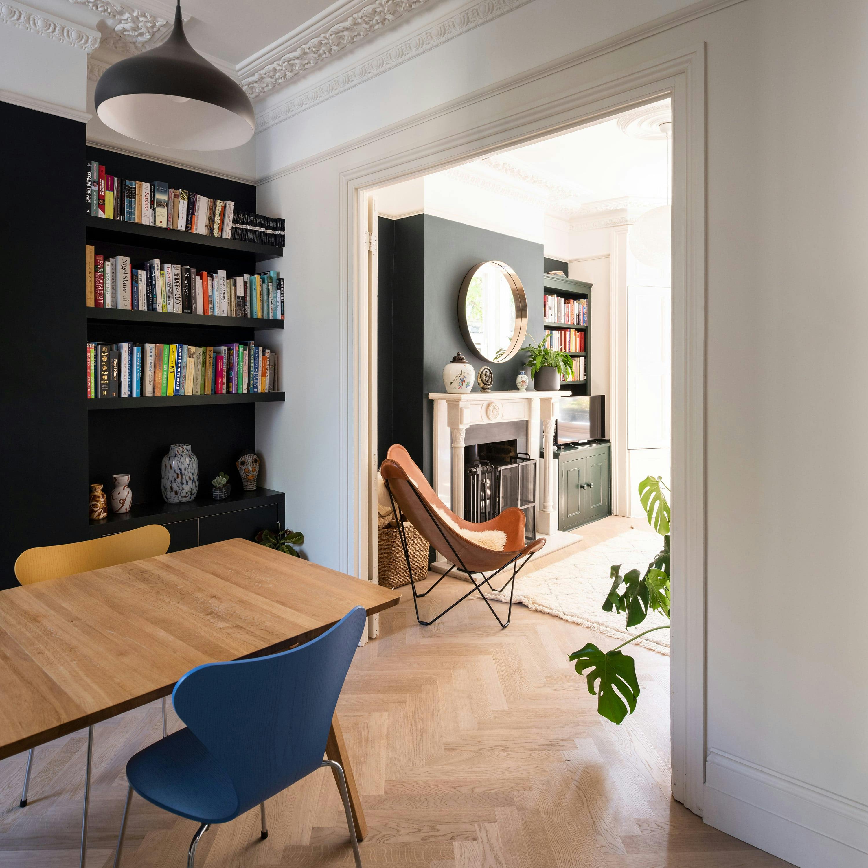 A stylish, modern interior of a townhouse featuring a dining area and a living room. The dining area has a wooden table with mismatched chairs in blue and yellow, set against a dark green built-in bookshelf filled with books and decorative objects. A black pendant light hangs from the ceiling. The living room, visible through an open doorway, has a classic white fireplace with a round mirror above it, flanked by bookshelves. A brown leather butterfly chair sits in front of the fireplace on a light-coloured rug. The space is illuminated by natural light, highlighting the intricate crown moulding and herringbone wood flooring. A potted monstera plant adds greenery to the scene.