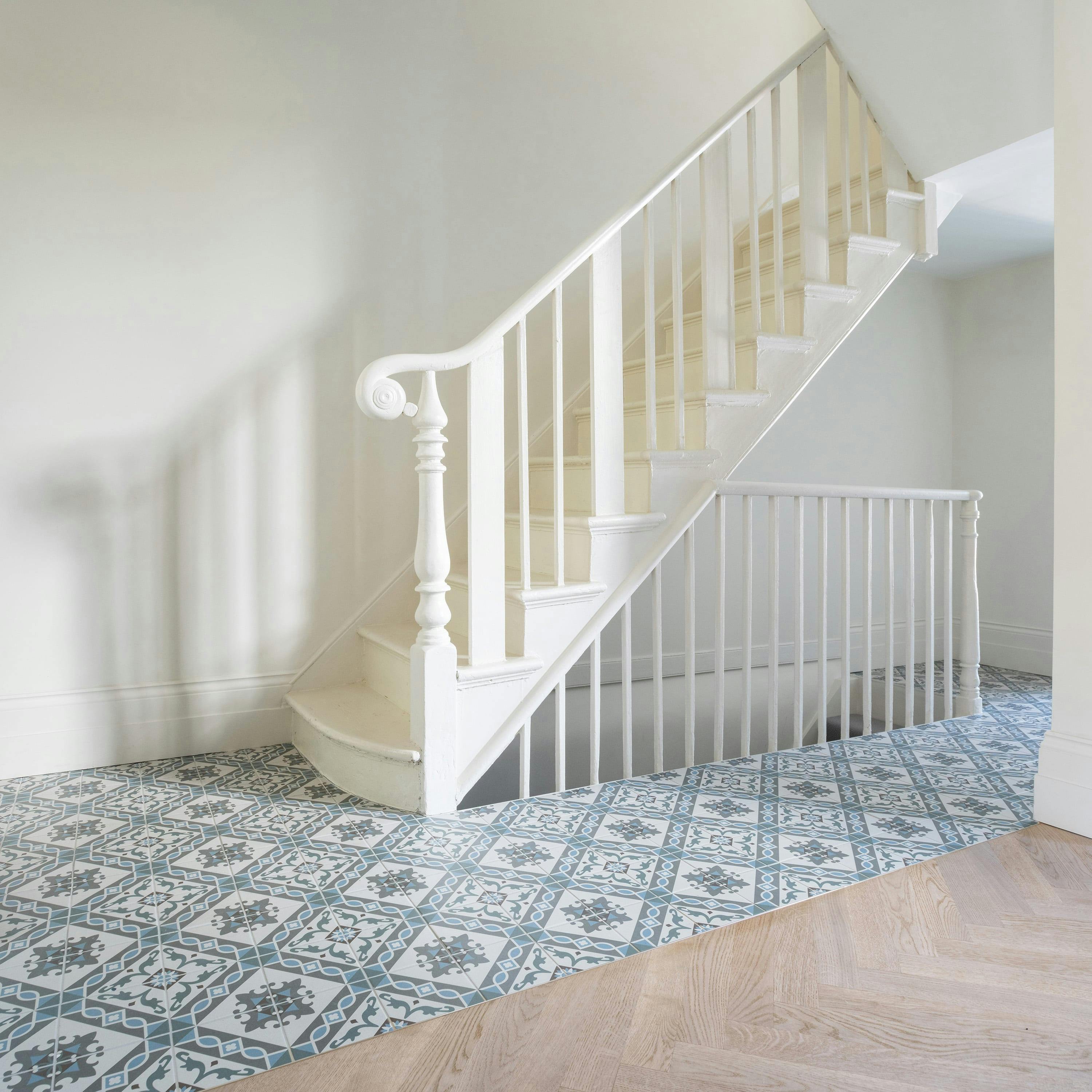 A bright and elegant hallway featuring a classic white wooden staircase with a curved handrail and vertical balusters. The lower steps have a rounded edge, adding a traditional touch. The flooring is a mix of patterned blue and white tiles near the staircase, transitioning into warm-toned herringbone wood flooring. The walls are painted in a soft, neutral shade, and natural light casts gentle shadows on the wall, enhancing the airy and serene ambiance of the space.