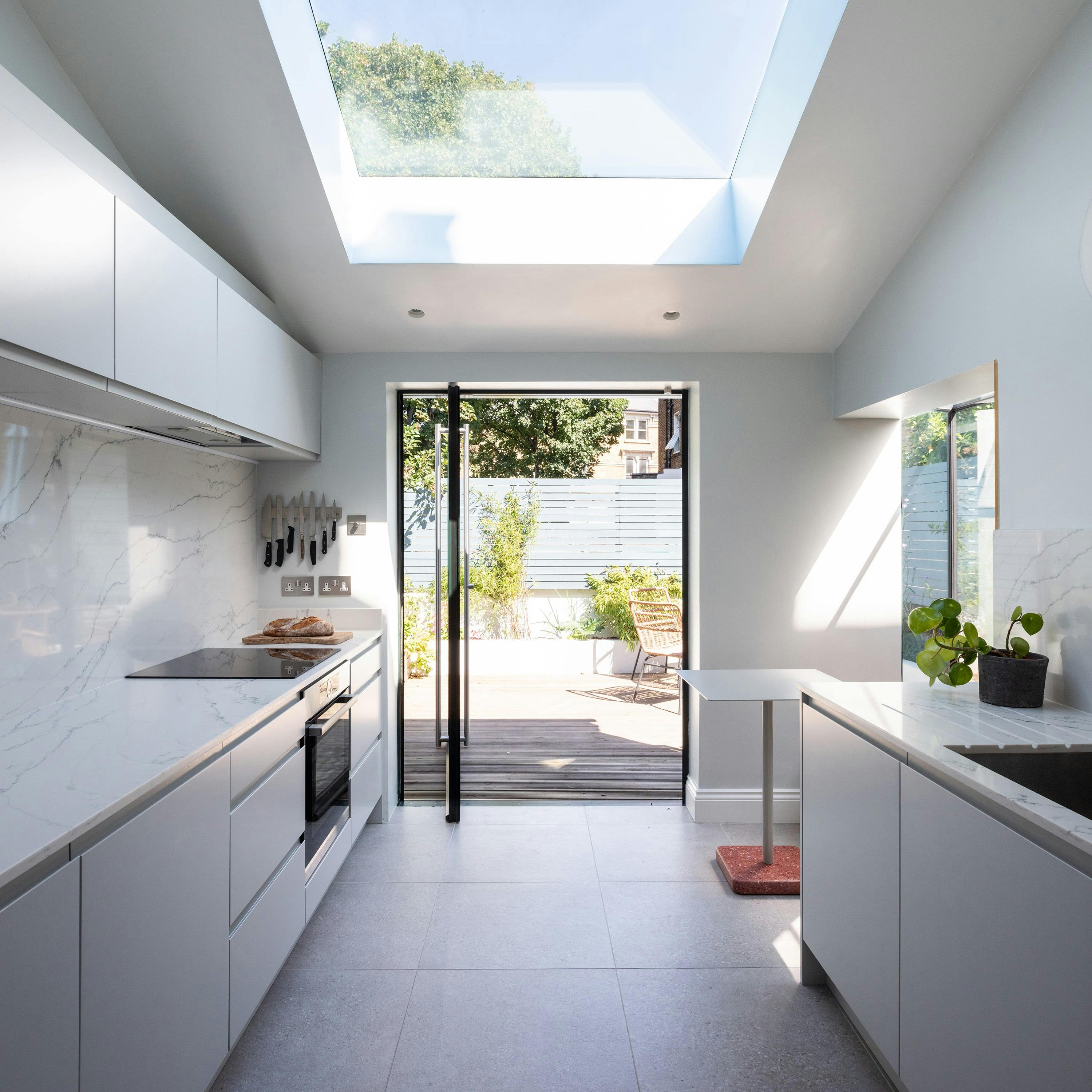 A modern, minimalist kitchen filled with natural light from a large skylight and glass doors that open to a lush outdoor patio. The kitchen features sleek white cabinetry, marble-effect countertops, and a backsplash. A built-in oven and induction cooktop are integrated into the cabinetry, with a magnetic knife rack mounted on the wall. On the right, a white countertop with a sink extends beneath a window, with a small potted plant adding a touch of greenery. The outdoor area is visible through the open doors, showing a wooden deck, seating, and a blue fence surrounded by greenery.