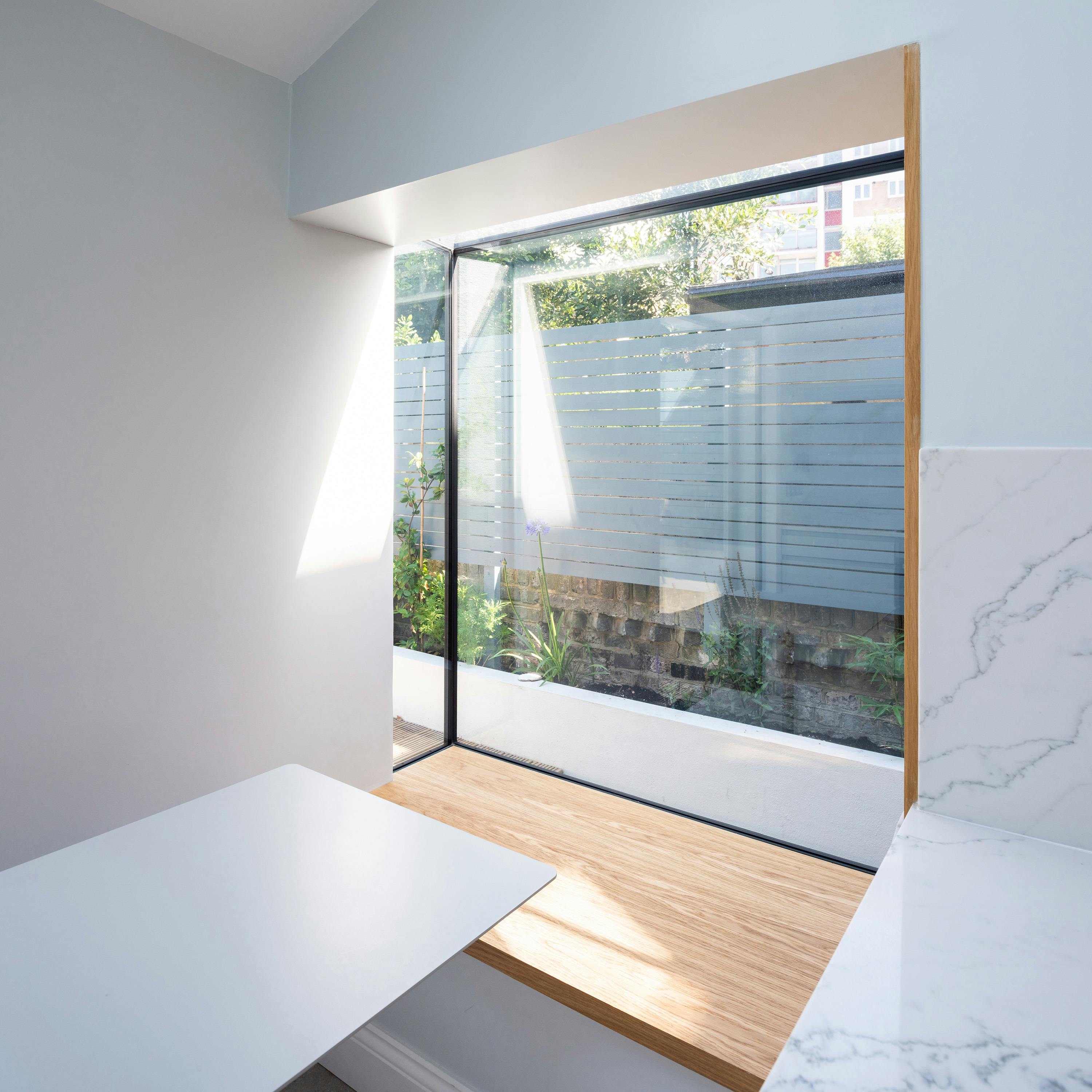 A modern kitchen nook featuring a large corner window with a wooden bench built into the frame. Natural light streams through the glass, illuminating the light-coloured walls and marble-effect countertops. The window offers a view of a small outdoor garden with plants and a blue slatted fence. A sleek white table extends from the wall, creating a functional dining or workspace. The design emphasizes minimalism, clean lines, and a seamless connection between the indoor and outdoor spaces.