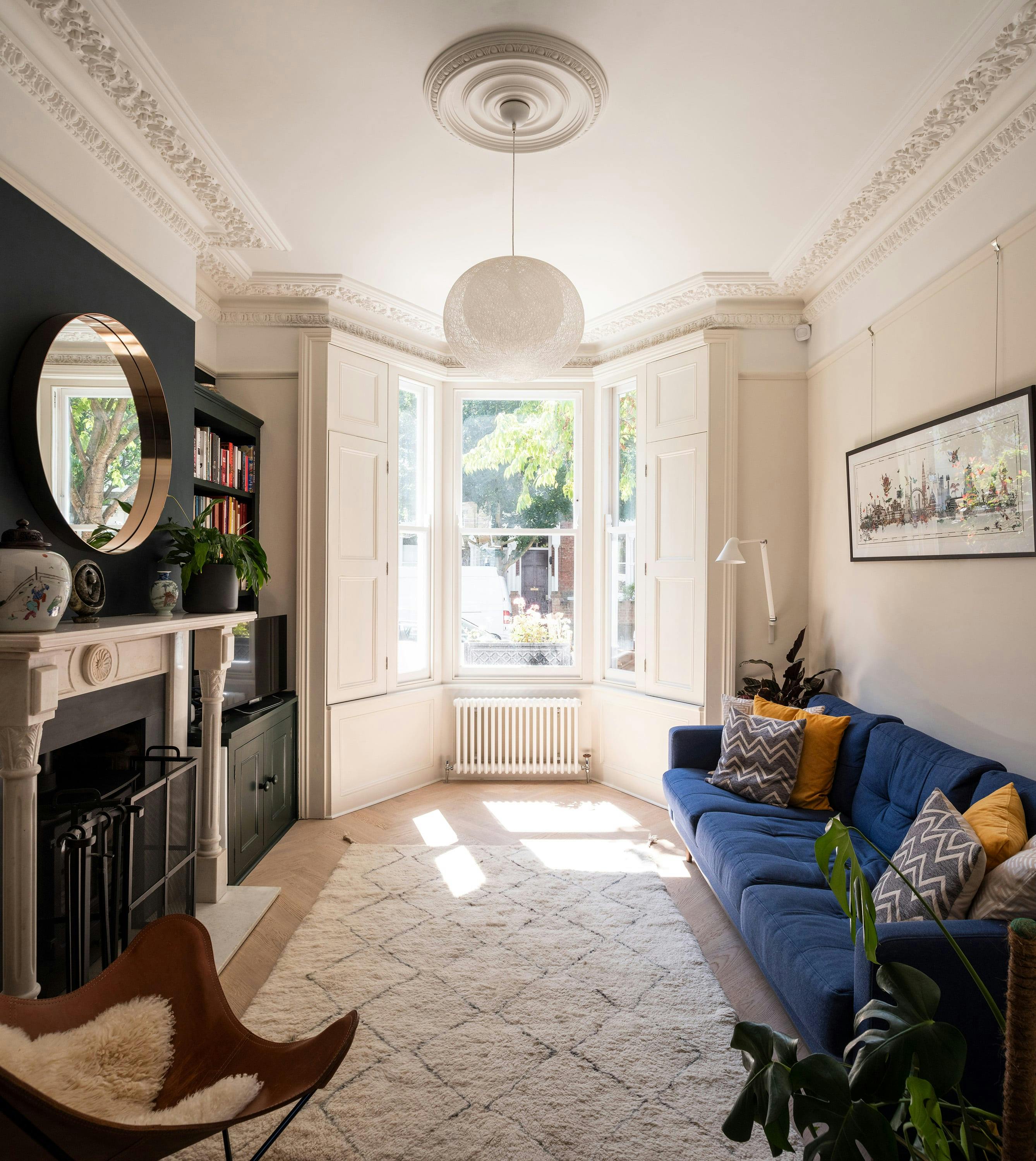 Elegant living room with a high ceiling, intricate crown moulding, and a large bay window allowing natural light to flood the space. A deep blue sofa with patterned and mustard-yellow throw pillows sits against a neutral-coloured wall, complemented by a framed artwork. A plush cream rug with a geometric pattern covers the wooden floor. The room features a classic white marble fireplace with a round mirror above it, built-in shelving filled with books, and potted plants adding greenery. A leather butterfly chair with a sheepskin throw sits in the foreground, enhancing the cozy yet sophisticated atmosphere.