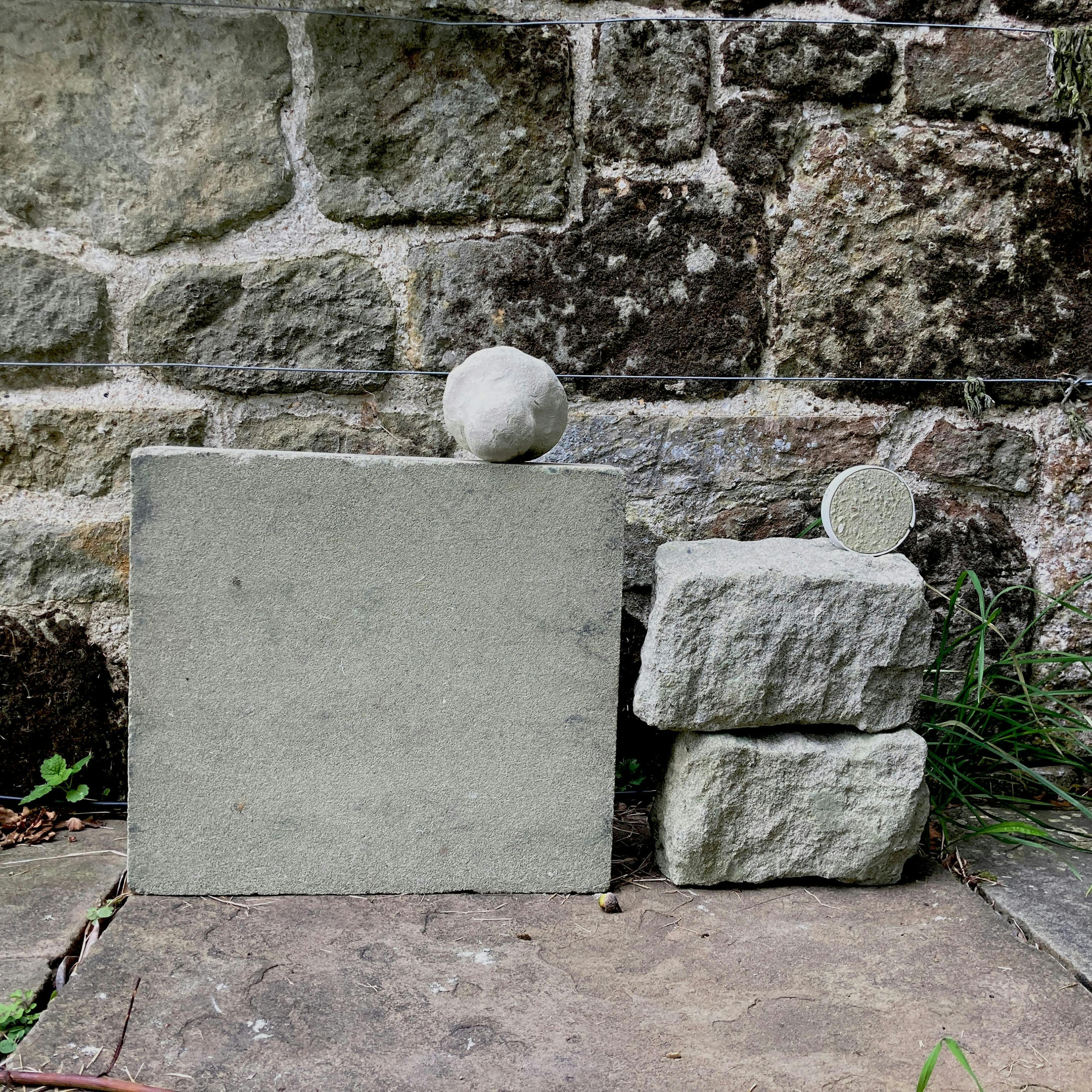 A square, smooth, light-colored stone slab sits next to two rougher, stacked stones of a similar color. A round, textured stone rests on top of the slab. A round, textured disc sits on top of the stacked stones. All are set against a weathered stone wall with visible mortar.