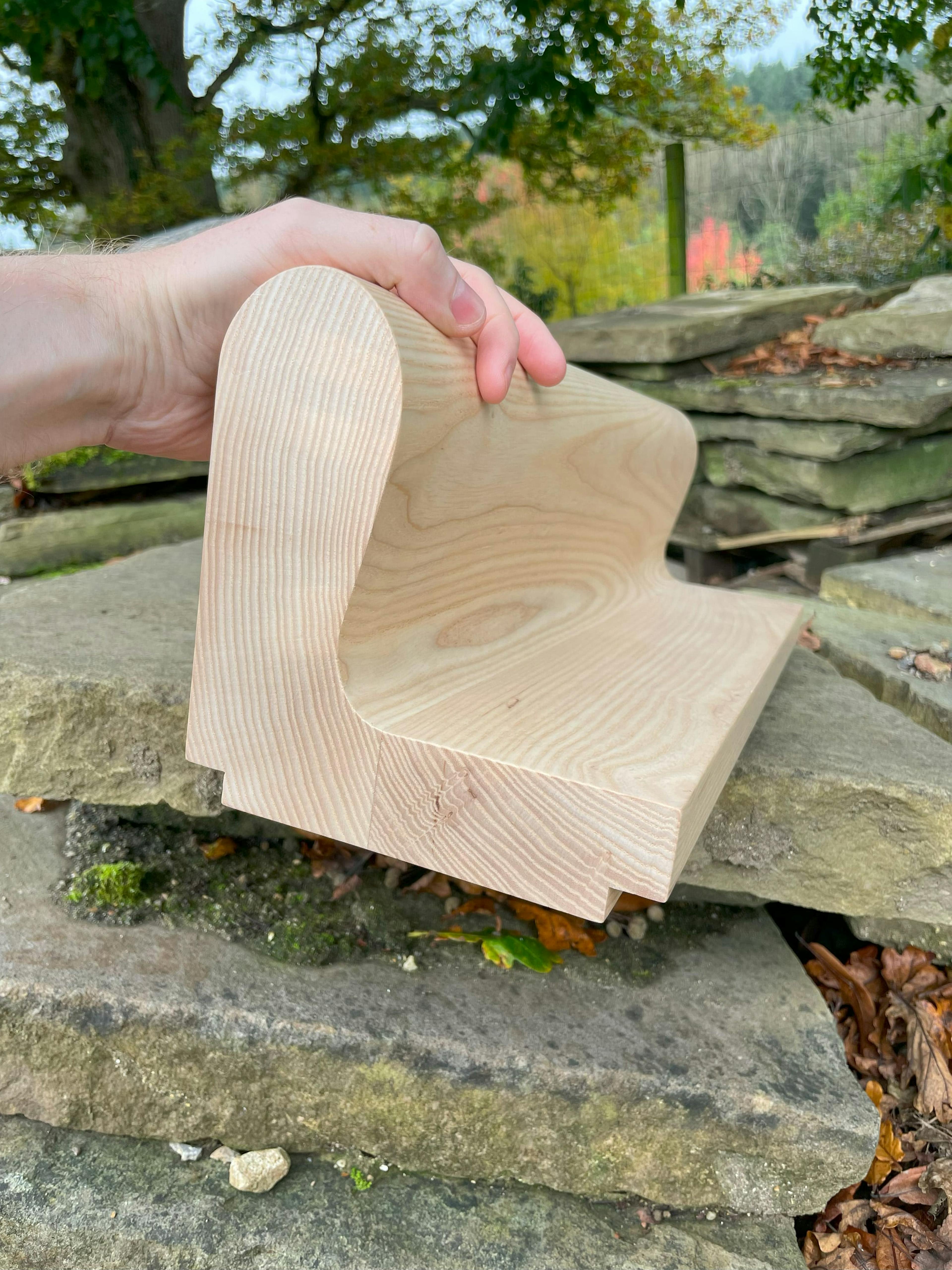A hand holds a light-colored wooden piece with a curved, contoured shape, possibly a section of a handrail. The wood grain is visible. The piece is held against a background of stacked, weathered stones and a blurred green landscape.