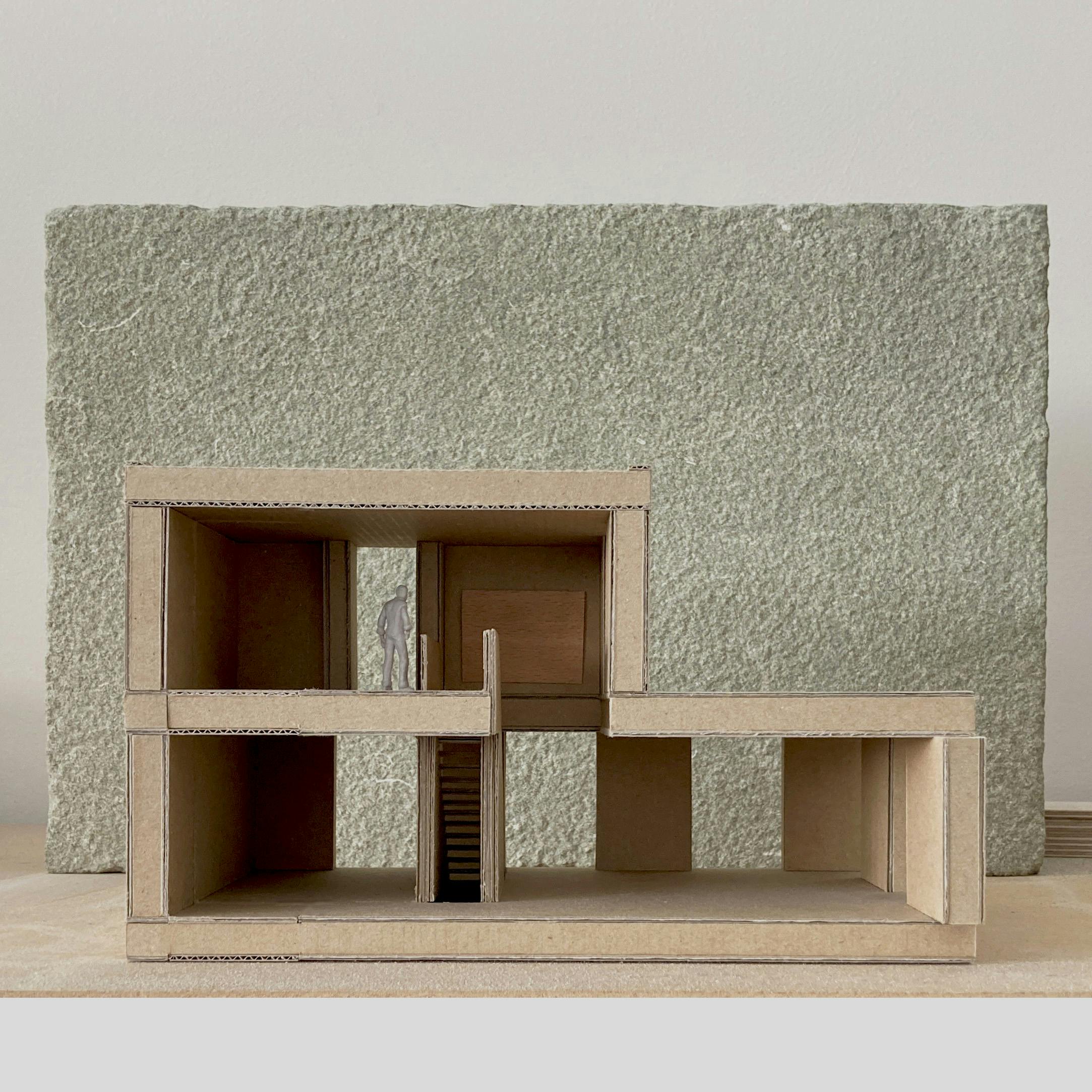 A cardboard architectural model of Casa G Escala is displayed with a textured stone slab as a backdrop. The model features open spaces, a staircase, and a figure inside. The model is made of corrugated cardboard.