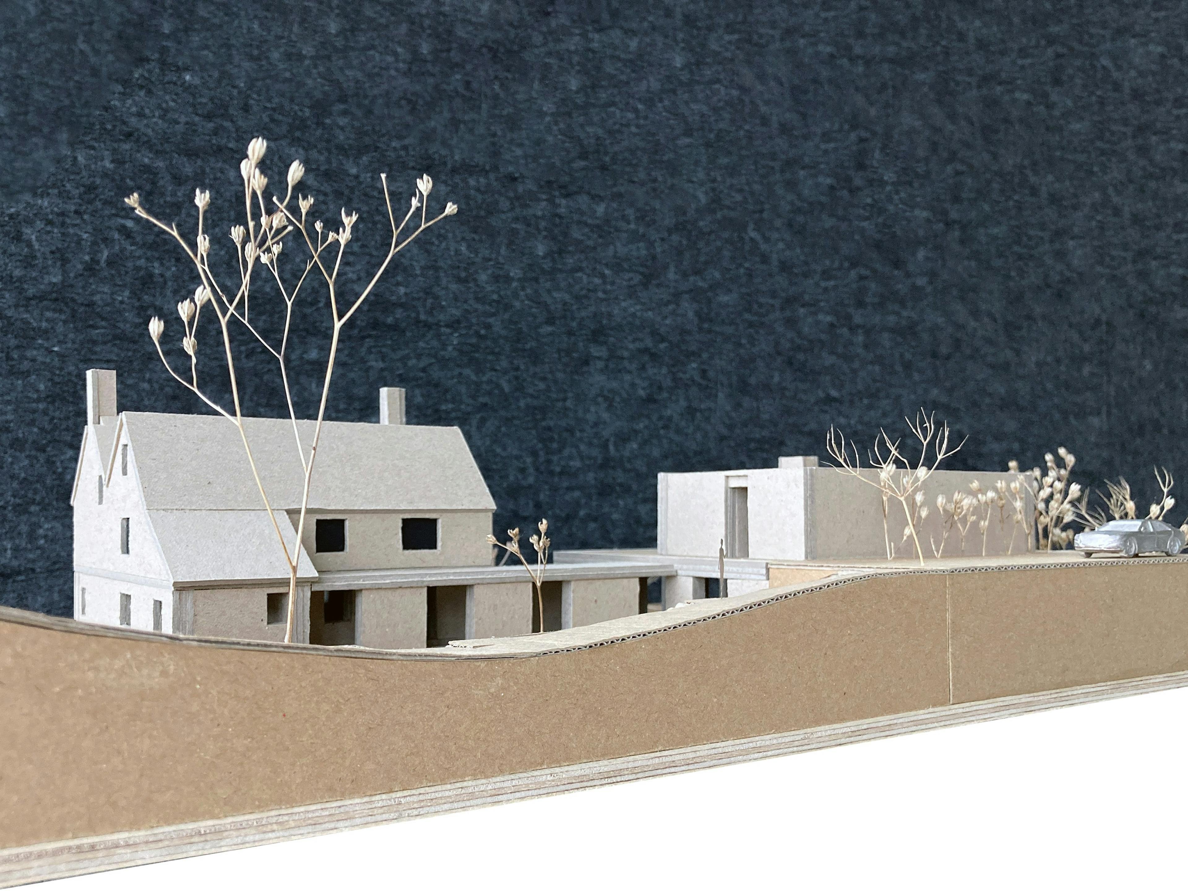A detailed cardboard architectural model showcases a house with a pitched roof and a modern building on a raised platform with a retaining wall. Dried plant stems are used to represent trees and foliage. A small car model is visible. The model has a dark grey textured backdrop.