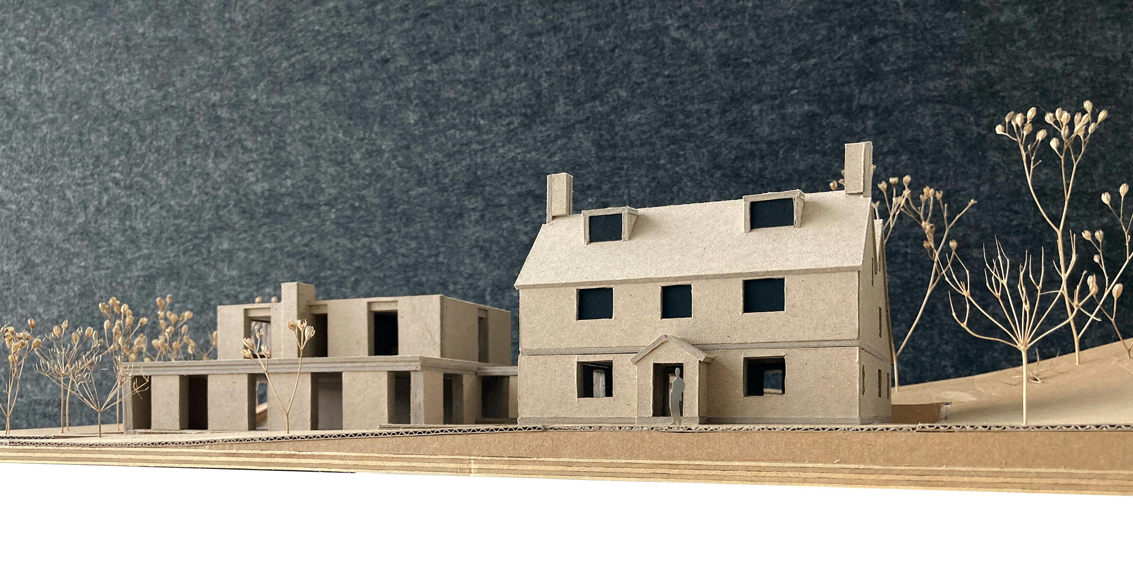 A detailed cardboard architectural model showcases a traditional house with a pitched roof and a contemporary building with flat roofs. Dried plant stems with seed heads are used to create landscape elements. The model has a dark grey textured backdrop.