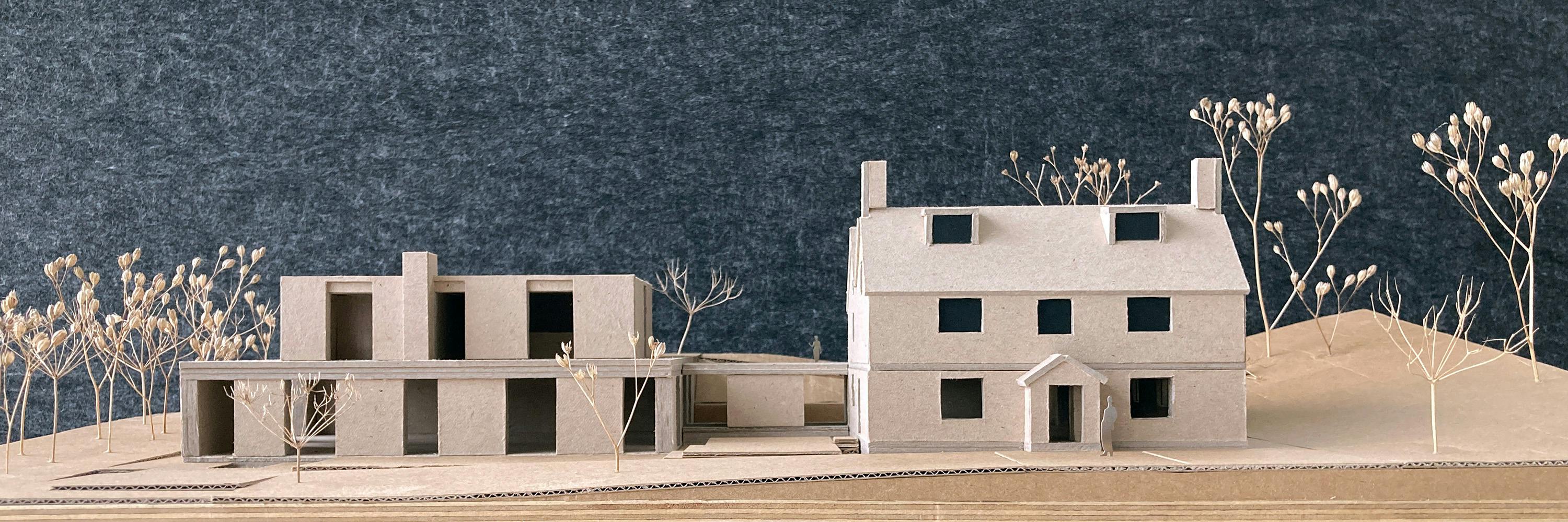 A cardboard architectural model depicts two buildings, including Greenstone House in Wiltshire, England: a house with a pitched roof and a modern building with flat roofs. Dried plant stems are used to represent trees and foliage. The model is set against a dark grey textured background.