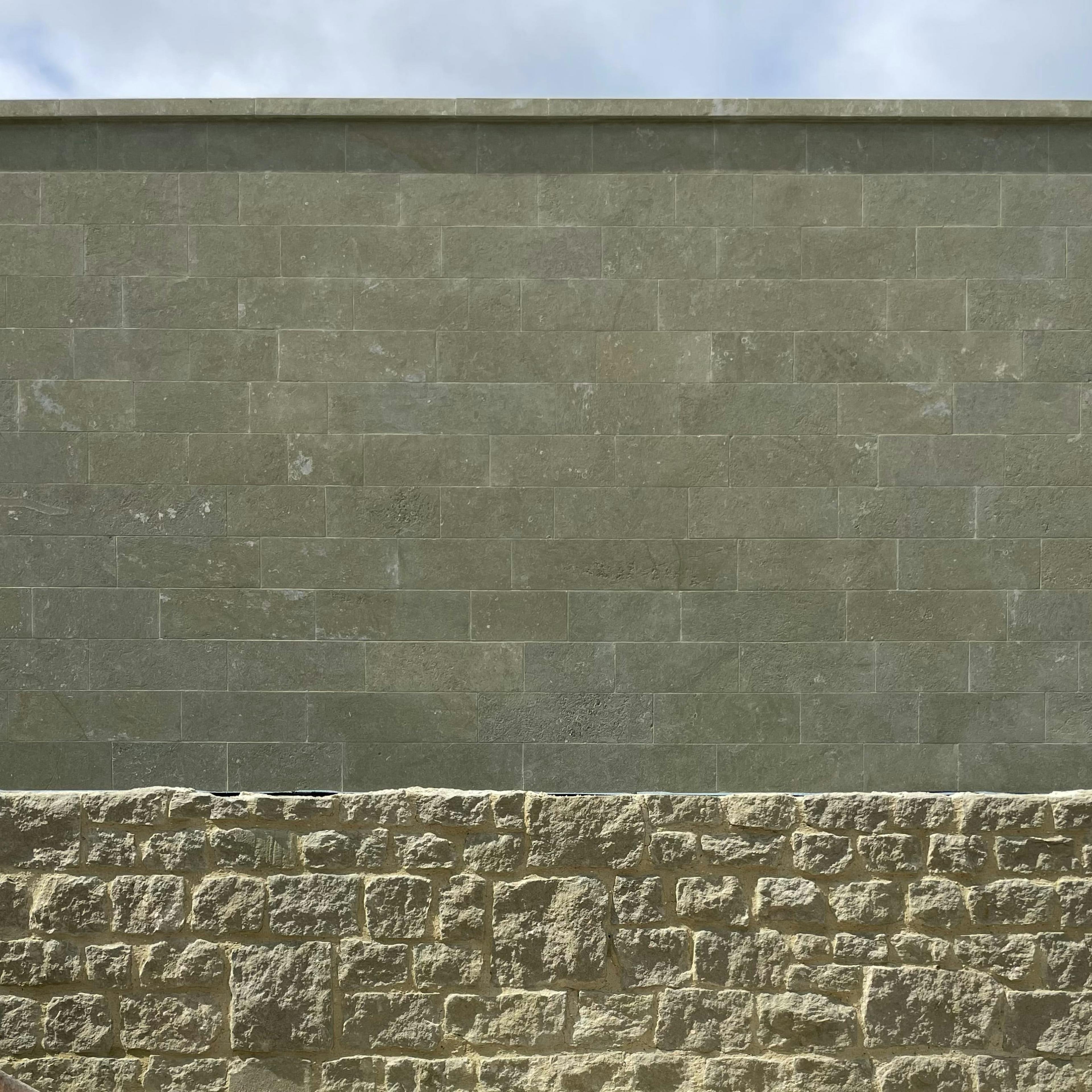 A wall with two distinct textures: the lower portion is made of rough, irregularly shaped beige stone blocks, while the upper portion consists of smooth, greenish rectangular tiles arranged in a uniform pattern. The sky with some clouds is visible above the wall.