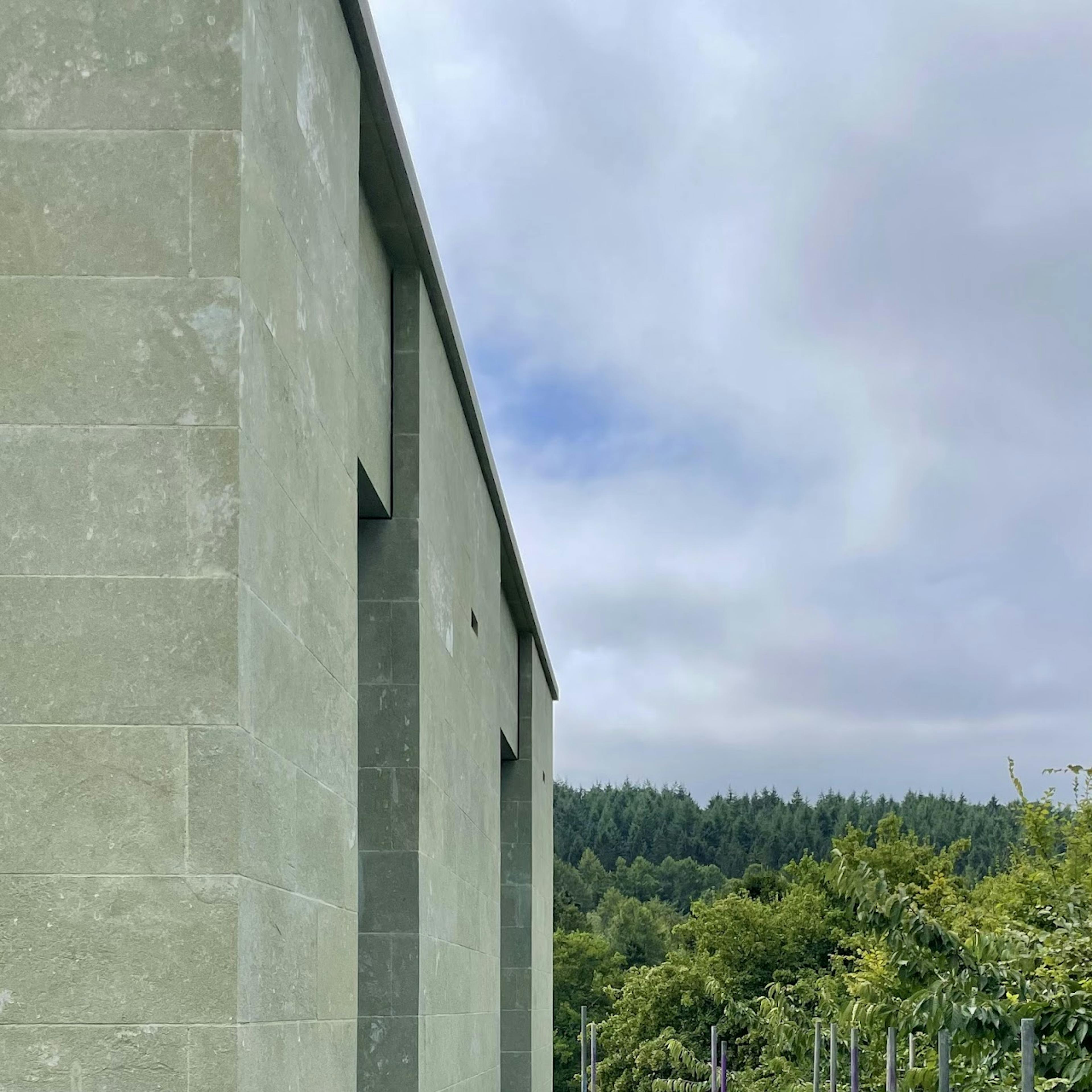 A modern building facade clad in greenish stone tiles with a smooth finish. The structure features recessed rectangular openings along its side. In the background, a lush green forest stretches across the horizon under a cloudy sky.
