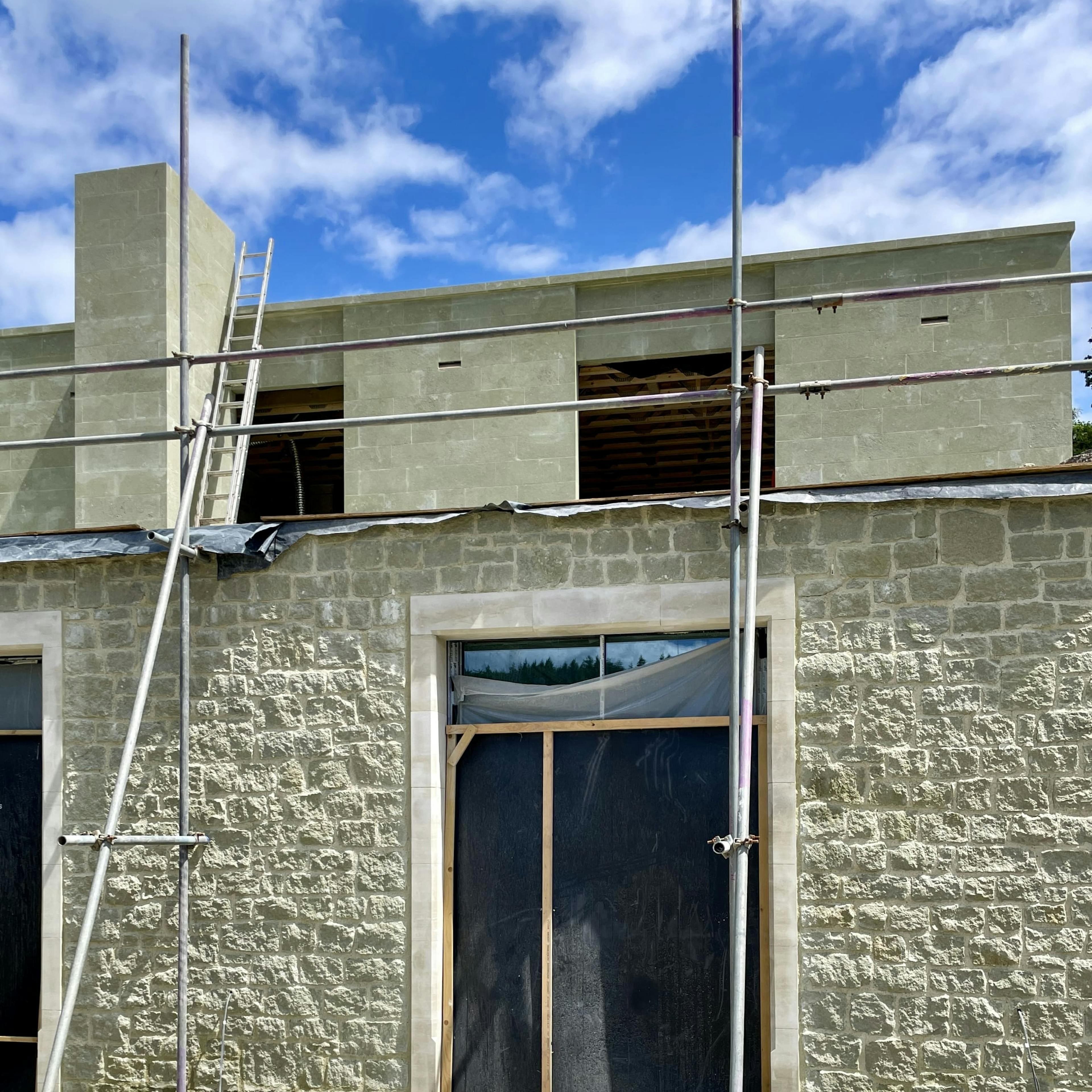 A building under construction with scaffolding in front. The lower portion of the structure is made of rough beige stone, while the upper section features smooth greenish stone cladding. A ladder leans against the scaffolding, and an unfinished window opening reveals the interior framework. The sky is bright blue with scattered white clouds.