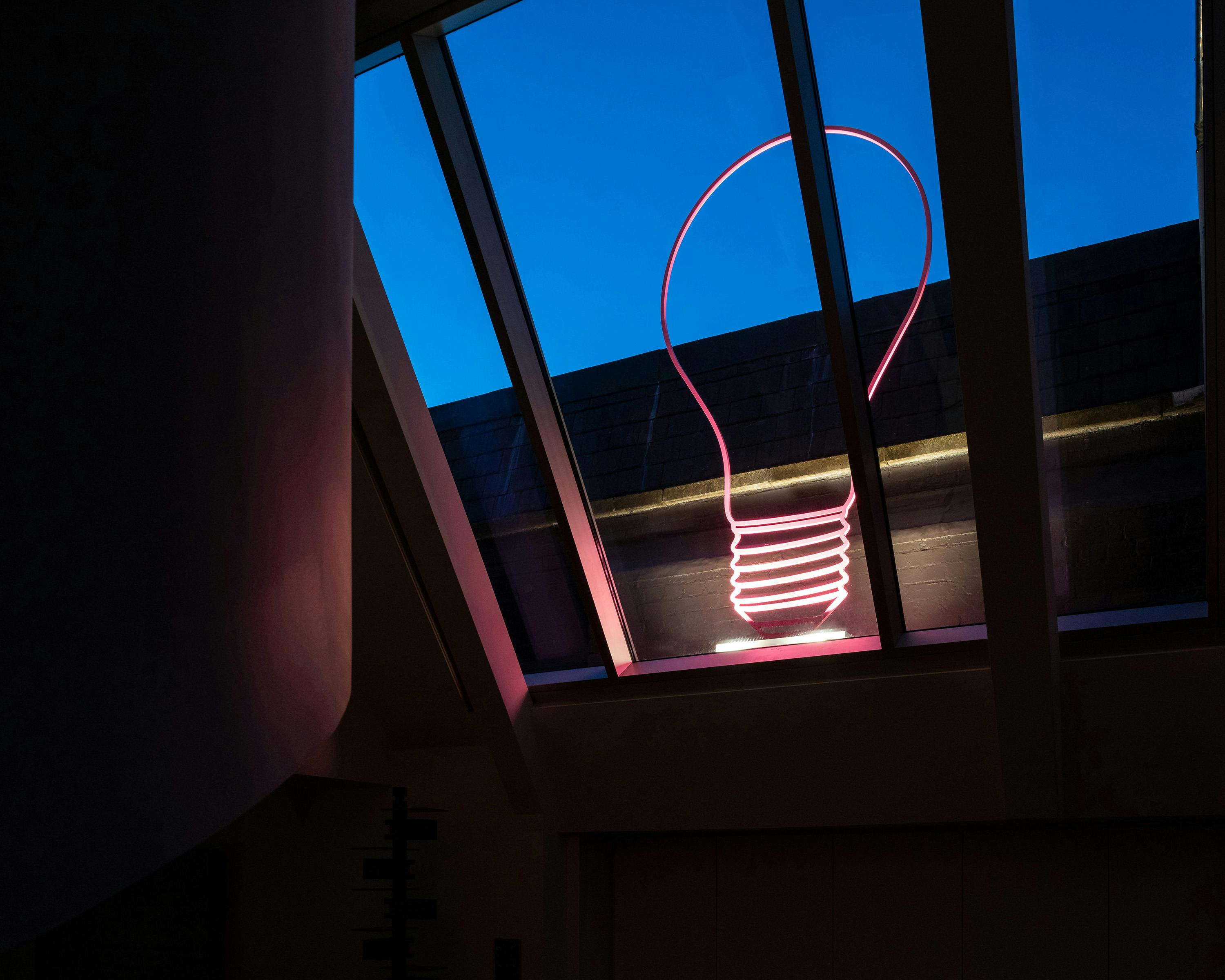 A pink neon lightbulb outline is visible in a window pane against a deep blue evening sky. The window is angled and framed by dark structural beams. The scene is dimly lit, suggesting dusk or dawn.