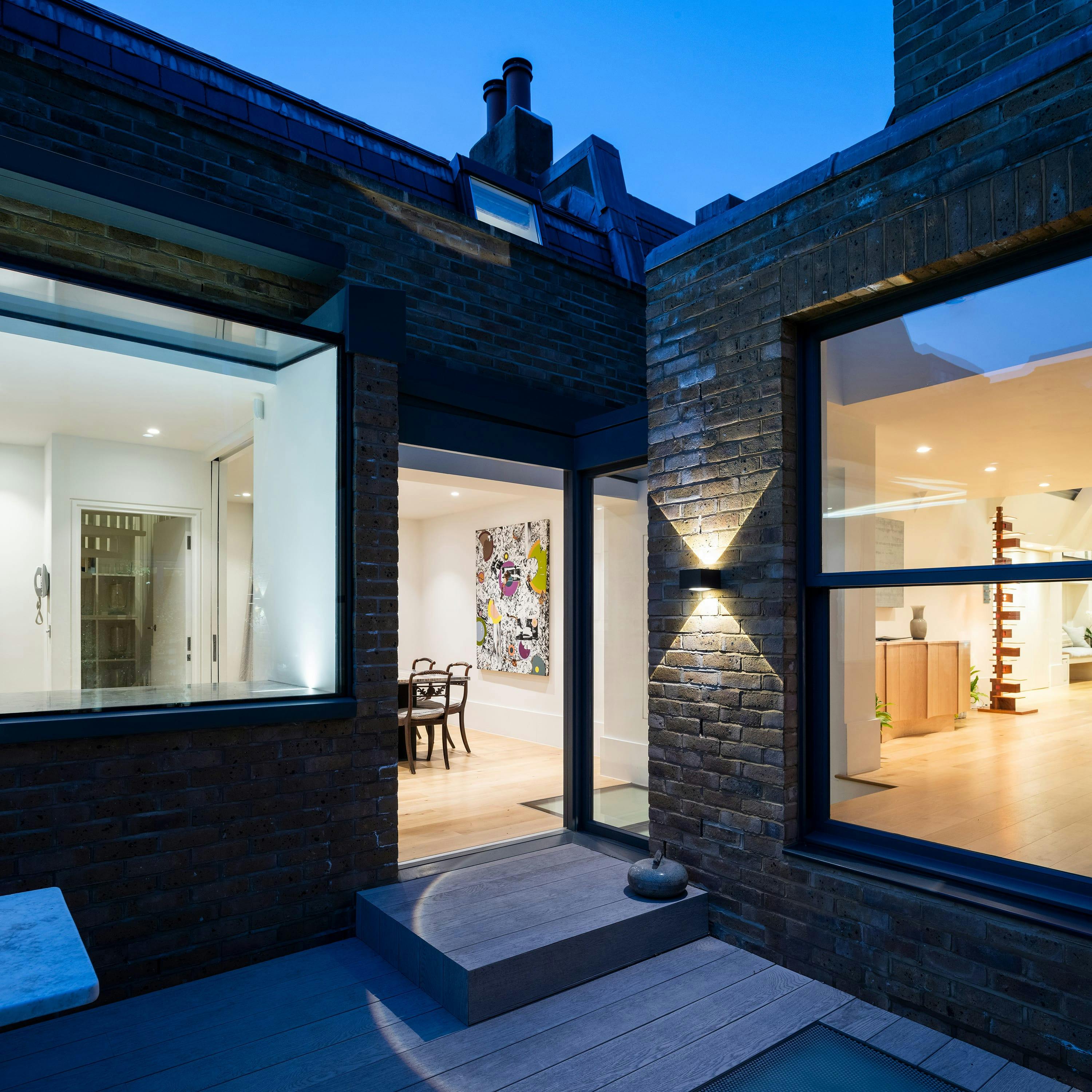 A modern architectural extension to a Chelsea townhouse is captured at dusk, showcasing a blend of traditional London architecture with contemporary design. The extension features dark brick, large windows revealing a dining space with artwork, and a paved courtyard with steps and decking. The sky is a deep twilight blue, highlighting the transition from day to night in this London neighborhood.