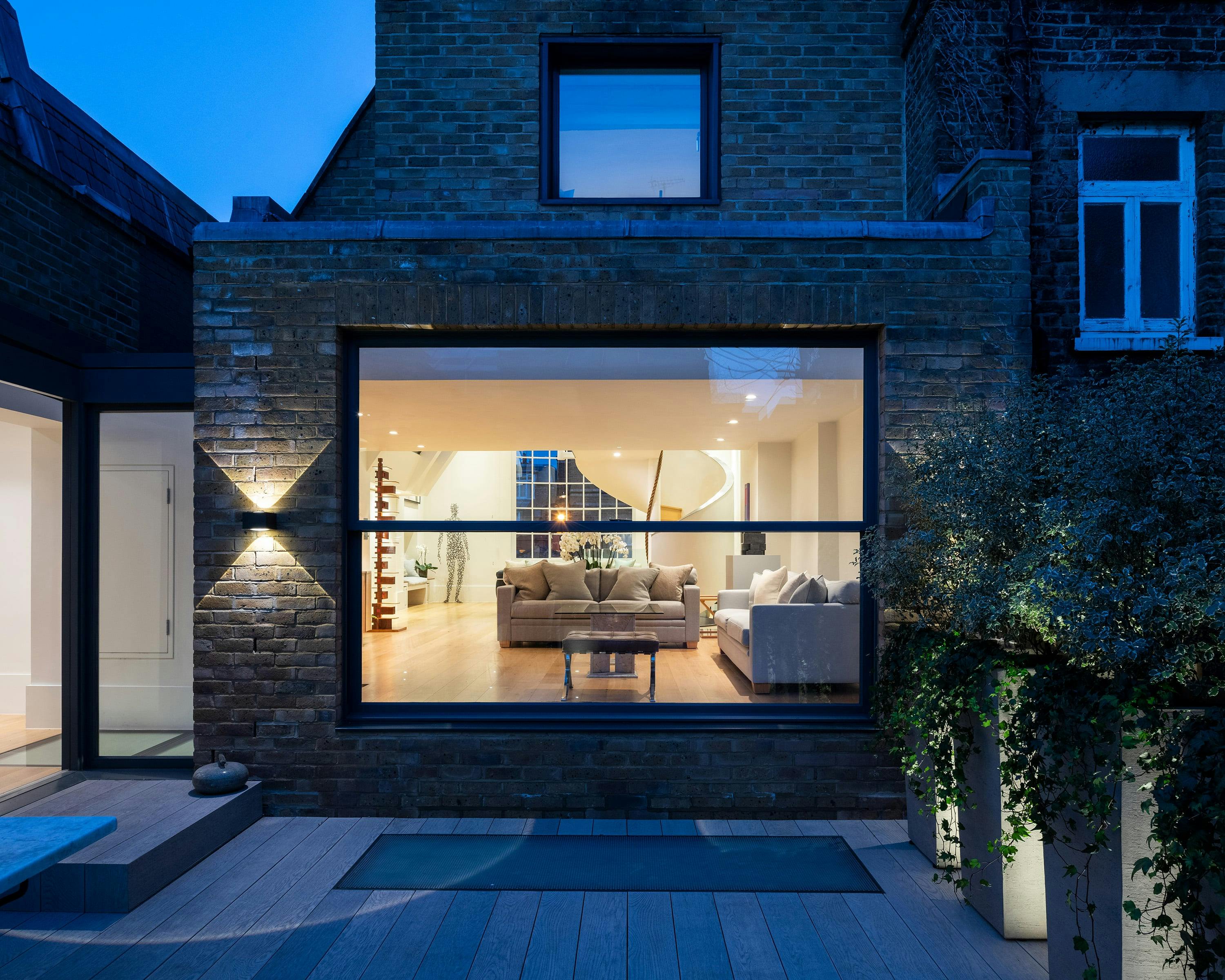 A modern architectural extension to a Chelsea townhouse is captured at night, showcasing a blend of traditional London architecture with contemporary design. The extension features dark brick, a large window revealing a well-lit living room with modern furniture, and a paved courtyard with decking and potted plants. The sky is dark, highlighting the contrast between the interior and exterior lighting.
