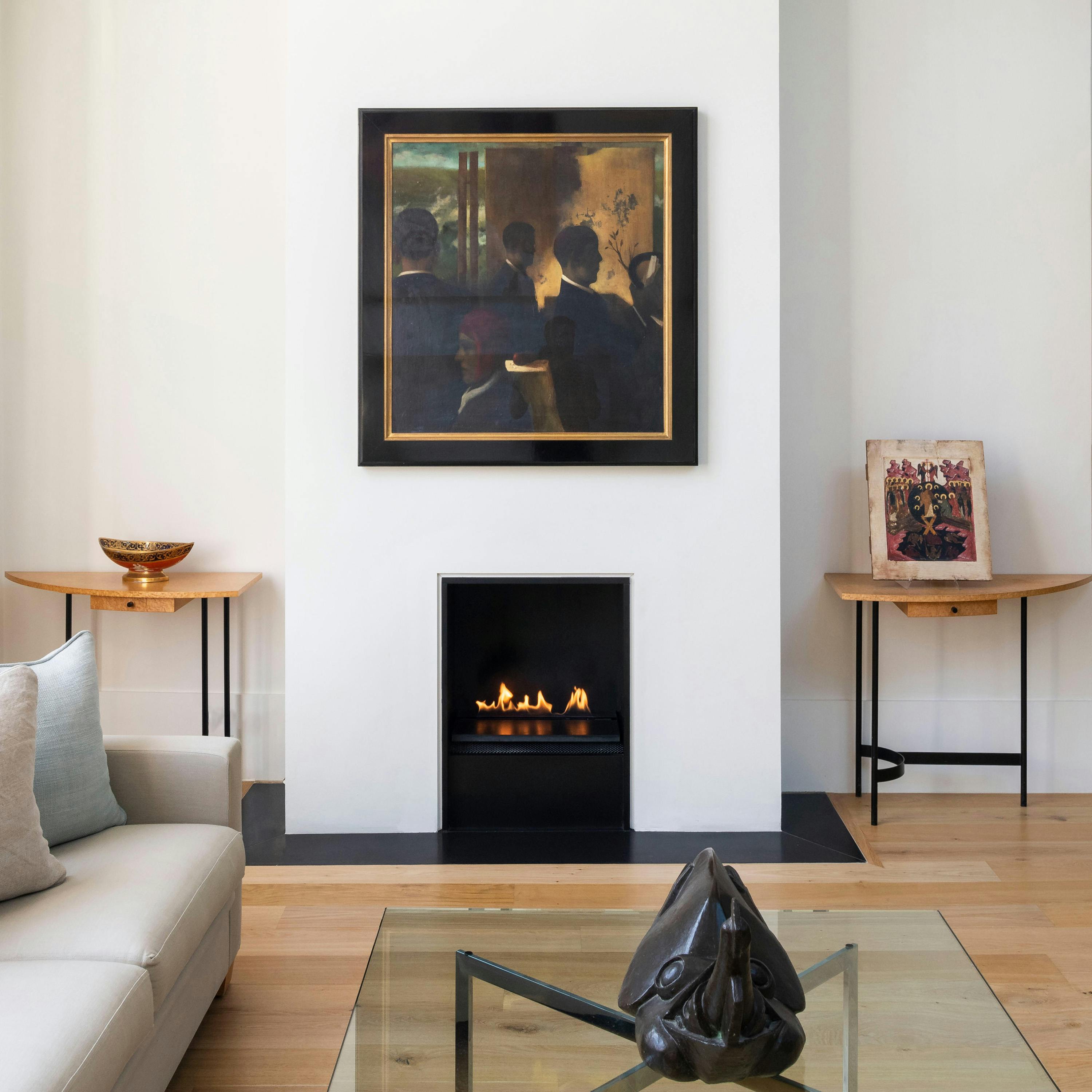 An interior shot of a contemporary living space with light wood flooring. A white wall features a framed painting with dark figures above a modern black fireplace with a gas flame. Two narrow wooden tables with metal legs are positioned on either side of the fireplace. A light-colored sofa is partially visible on the left, and a glass coffee table with a sculptural object is in the foreground.