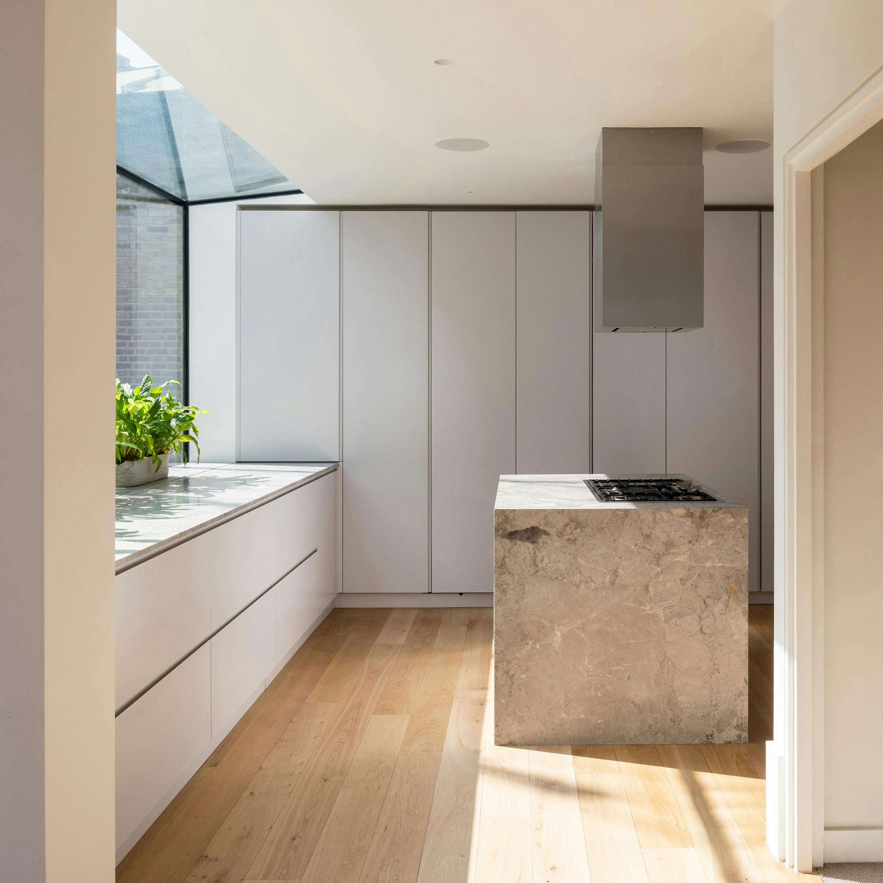 An interior shot of a contemporary kitchen with light wood floors and sleek, white cabinets. A large stone kitchen island with a built-in cooktop is the focal point. A skylight provides natural light. A potted plant adds a touch of greenery to the space.